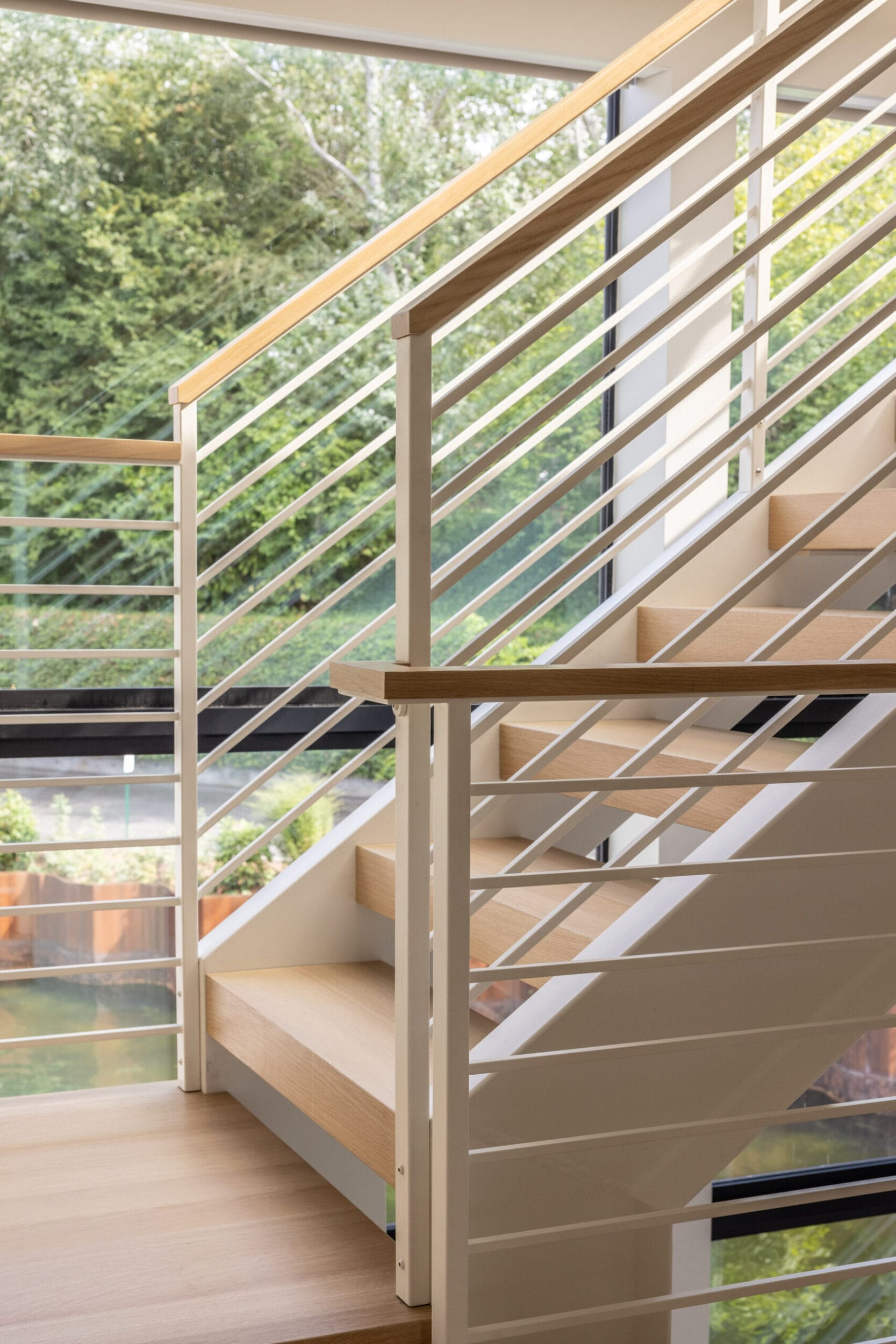 Modern indoor staircase with light wood steps and white metal railings, adjacent to large windows overlooking a green garden.