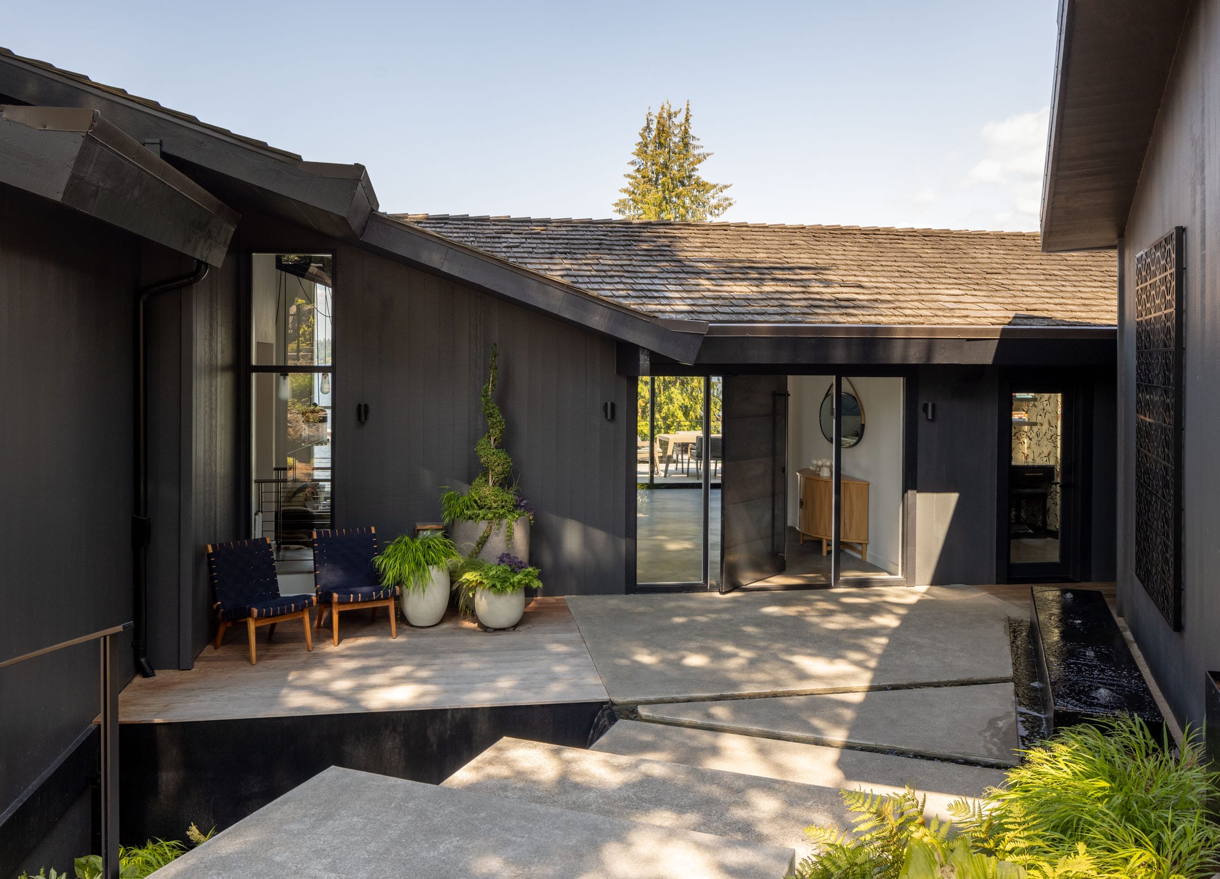 Modern house exterior with angular design, dark wood siding, large windows, and a sloped roof. Outdoor seating and planters with greenery enhance the entrance area.