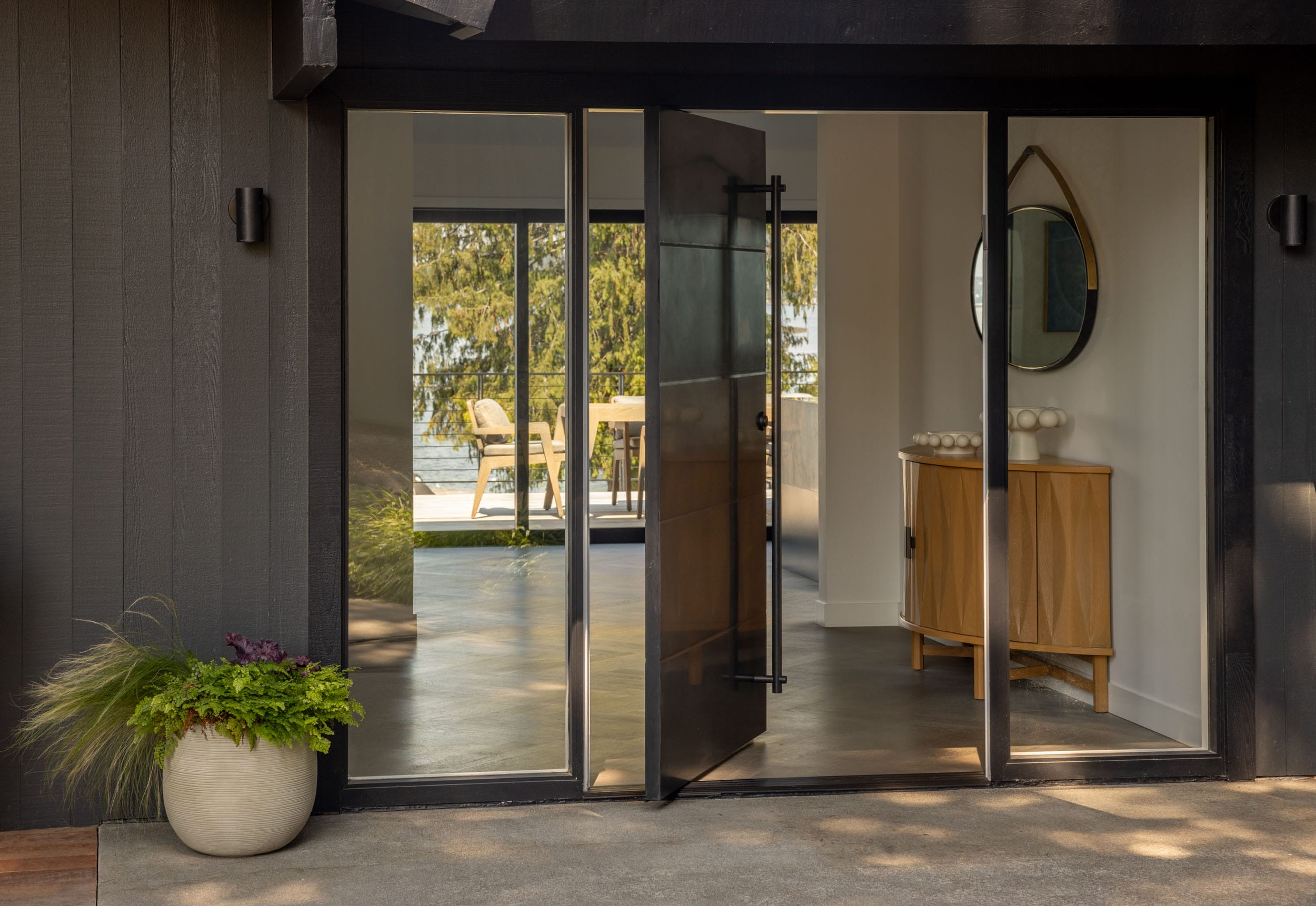 Modern entry with a black-framed glass door open to an interior, featuring a wooden cabinet and round mirror. Outdoor view reflects on the glass.