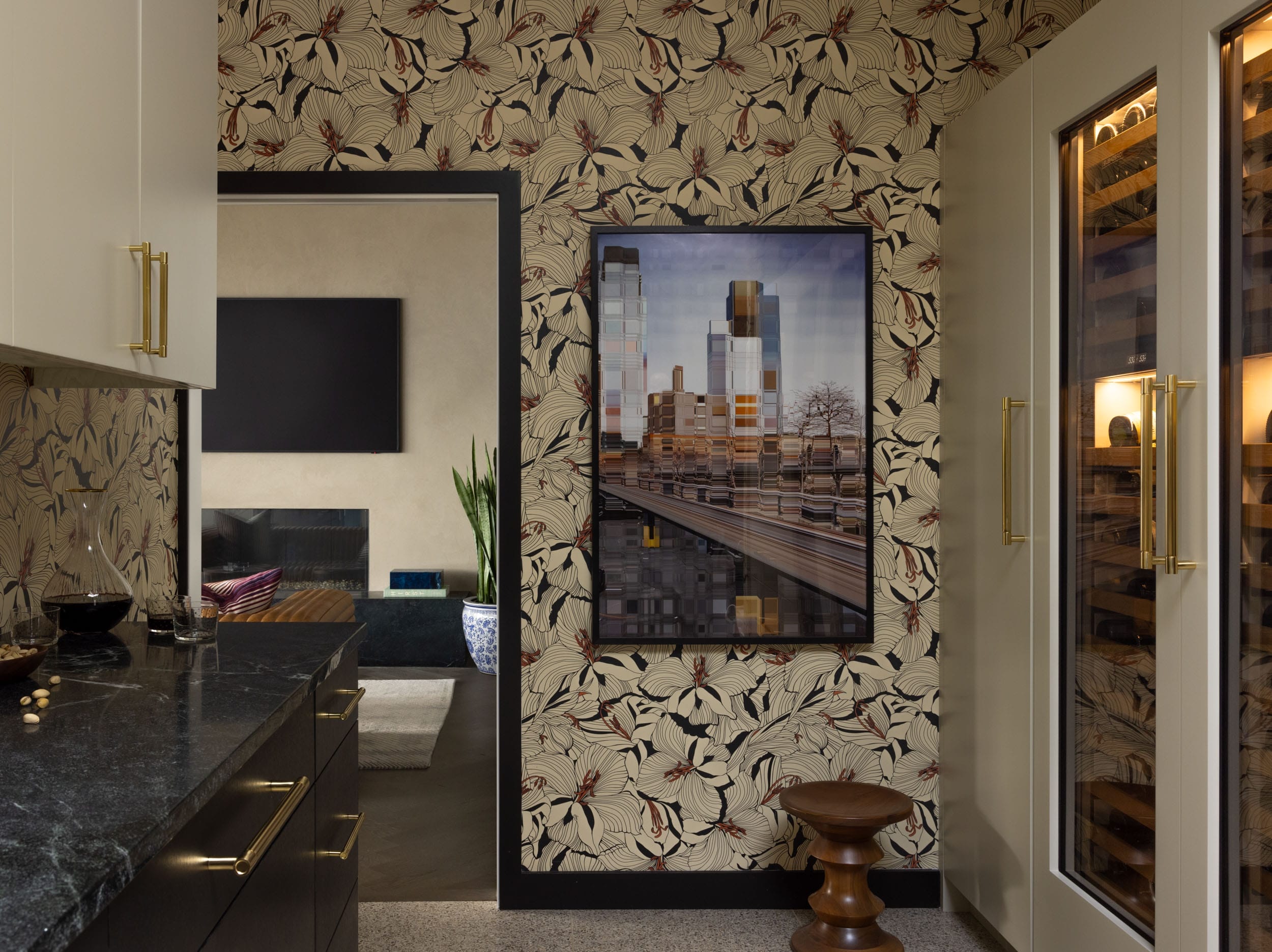 Stylish kitchen with floral wallpaper, dark countertops, a large framed cityscape photo, and a wine fridge with glass doors on the right. A glimpse of the adjacent living room is visible.