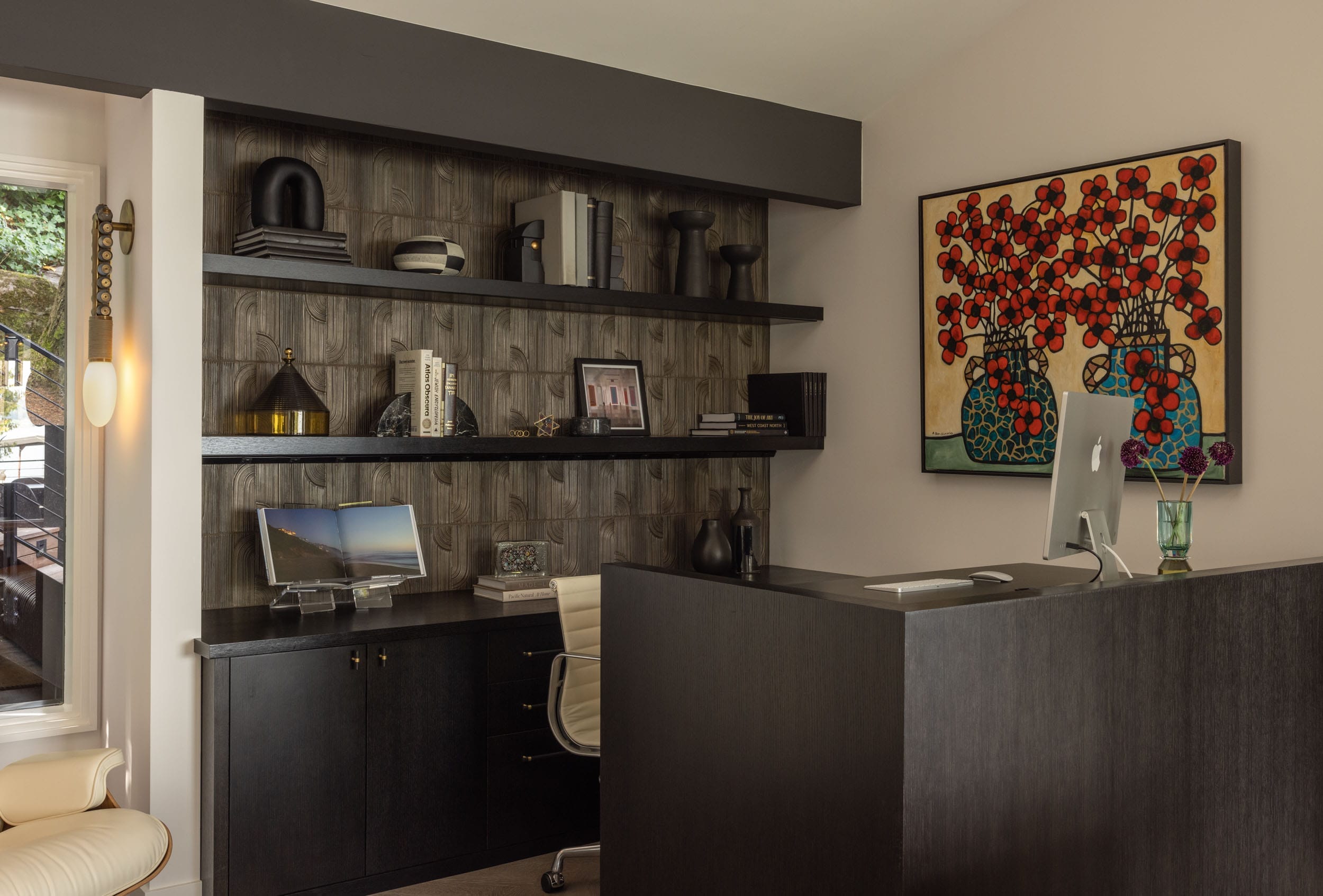 Home office with dark wood desk, shelves with books and decor, a window with a wall light, and a colorful floral painting on the wall.