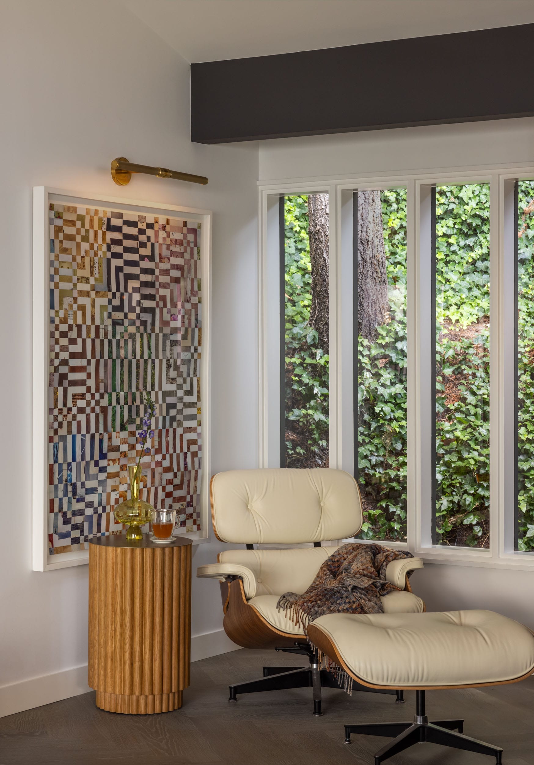 A cozy corner with an Eames lounge chair and ottoman, a wooden side table with a small plant, and a colorful geometric artwork on the wall near large windows with a leafy view.