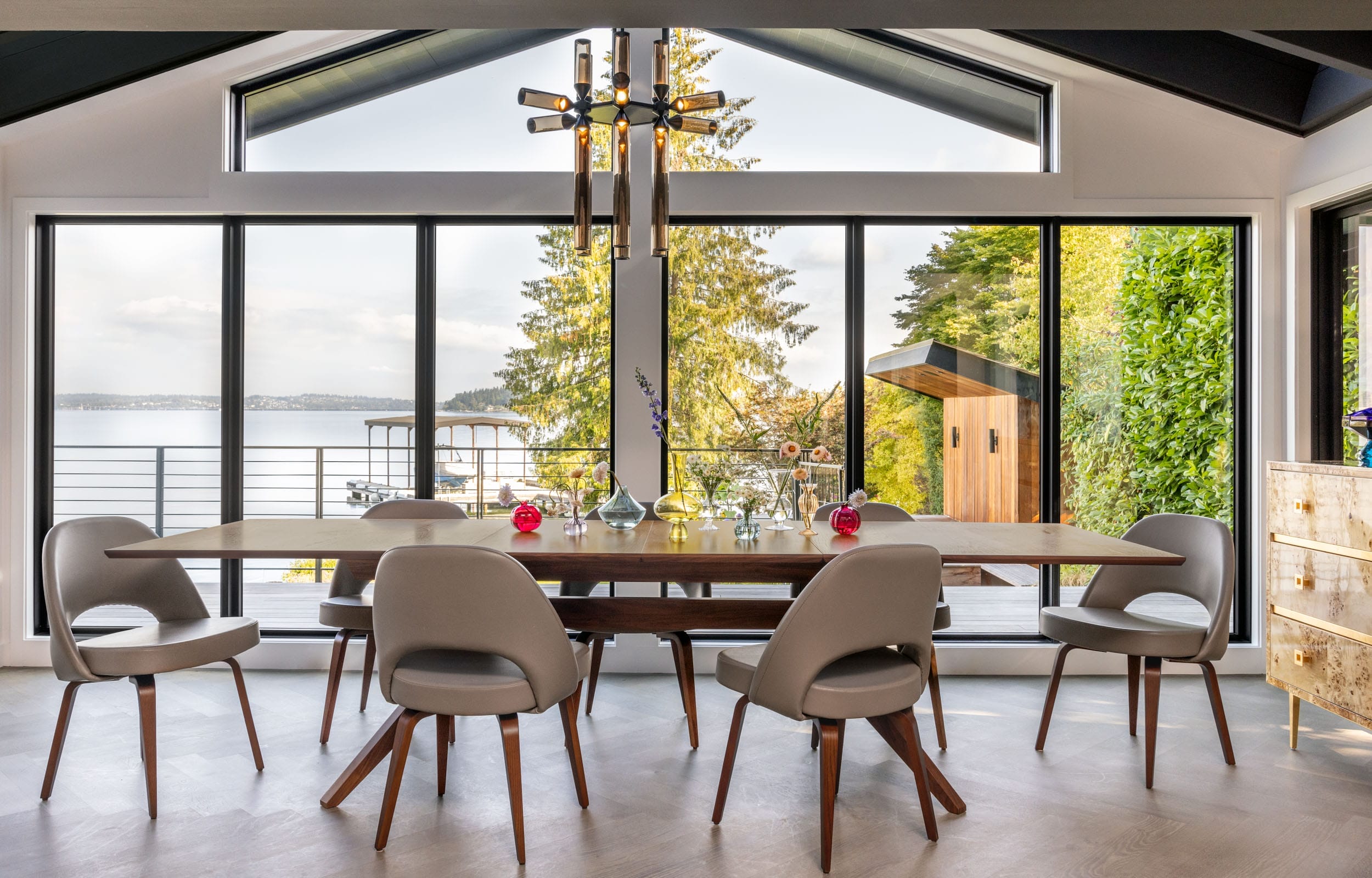 Modern dining room with a wooden table, six gray chairs, and a chandelier, overlooking a scenic lake view through large windows.