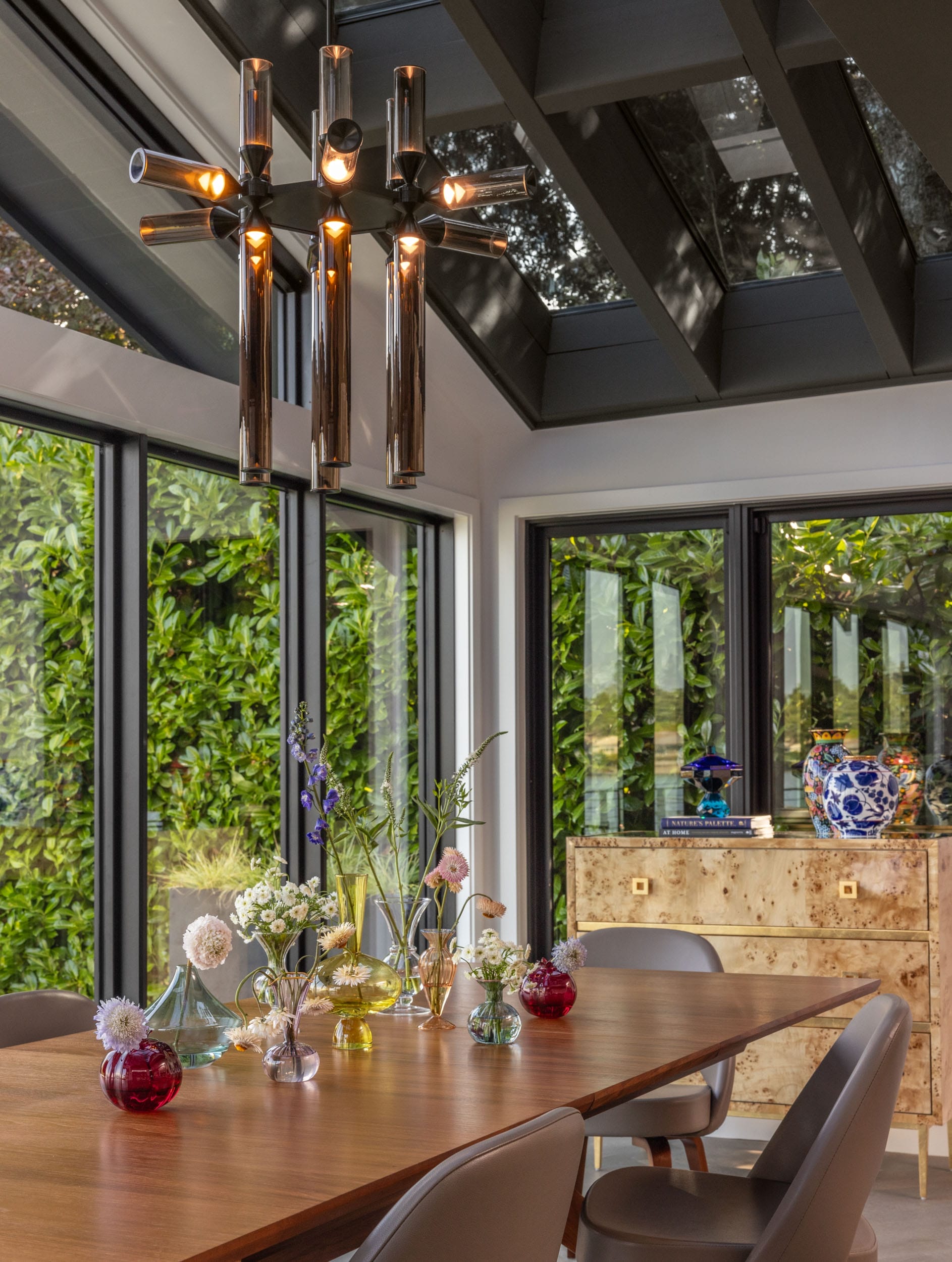 A dining room with a wooden table, modern chandelier, and decorative vases containing flowers. Large windows surround the room, showing greenery outside. A cabinet with a vase is in the corner.