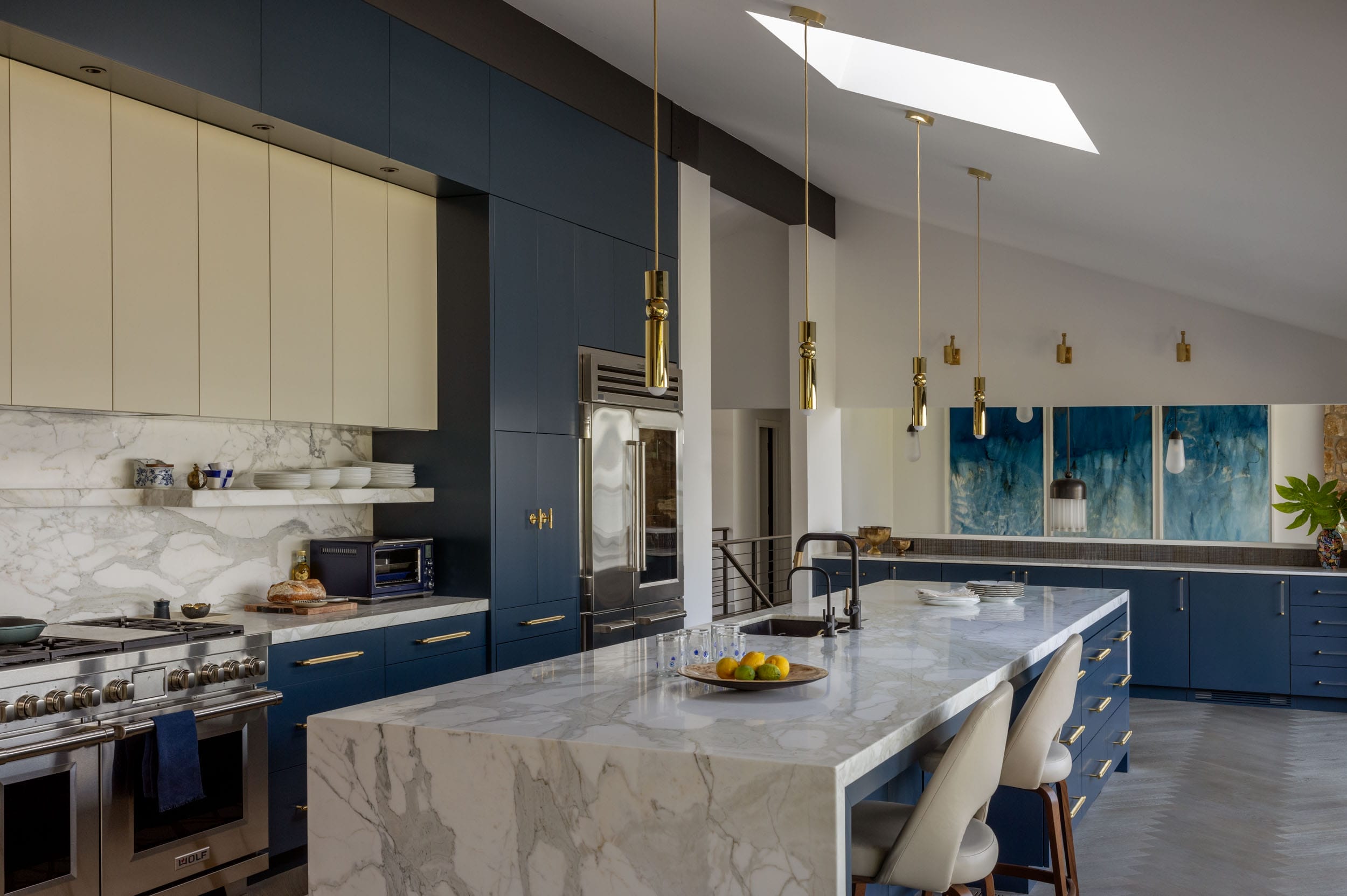 Modern kitchen with blue cabinets, marble countertops, and gold accents. Includes a large island with bar stools, pendant lighting, and a skylight above.