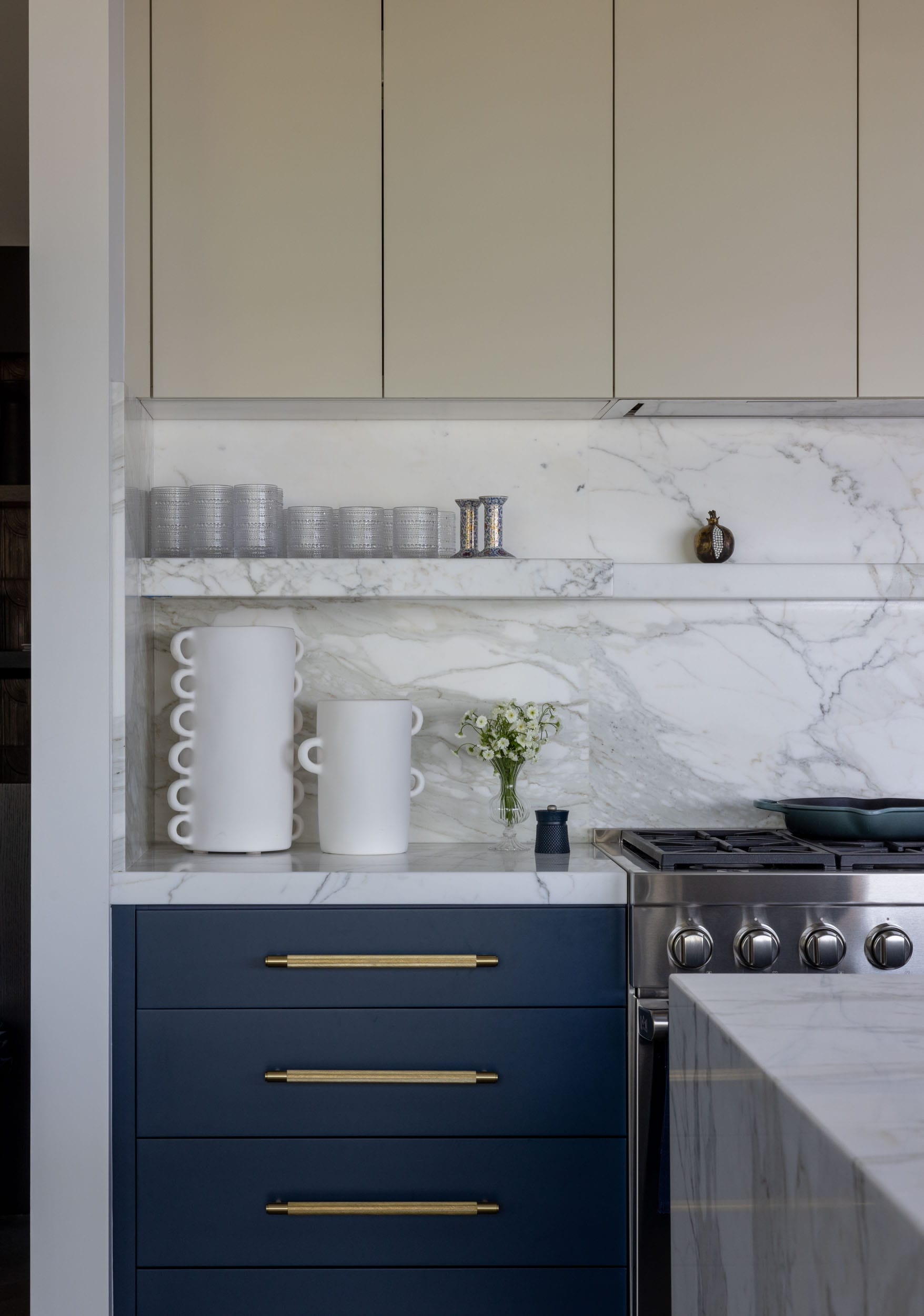 Modern kitchen with blue lower cabinets, a marble backsplash, and white upper cabinets. It features a stainless steel stove, white vases, stacked bowls, and a small plant.