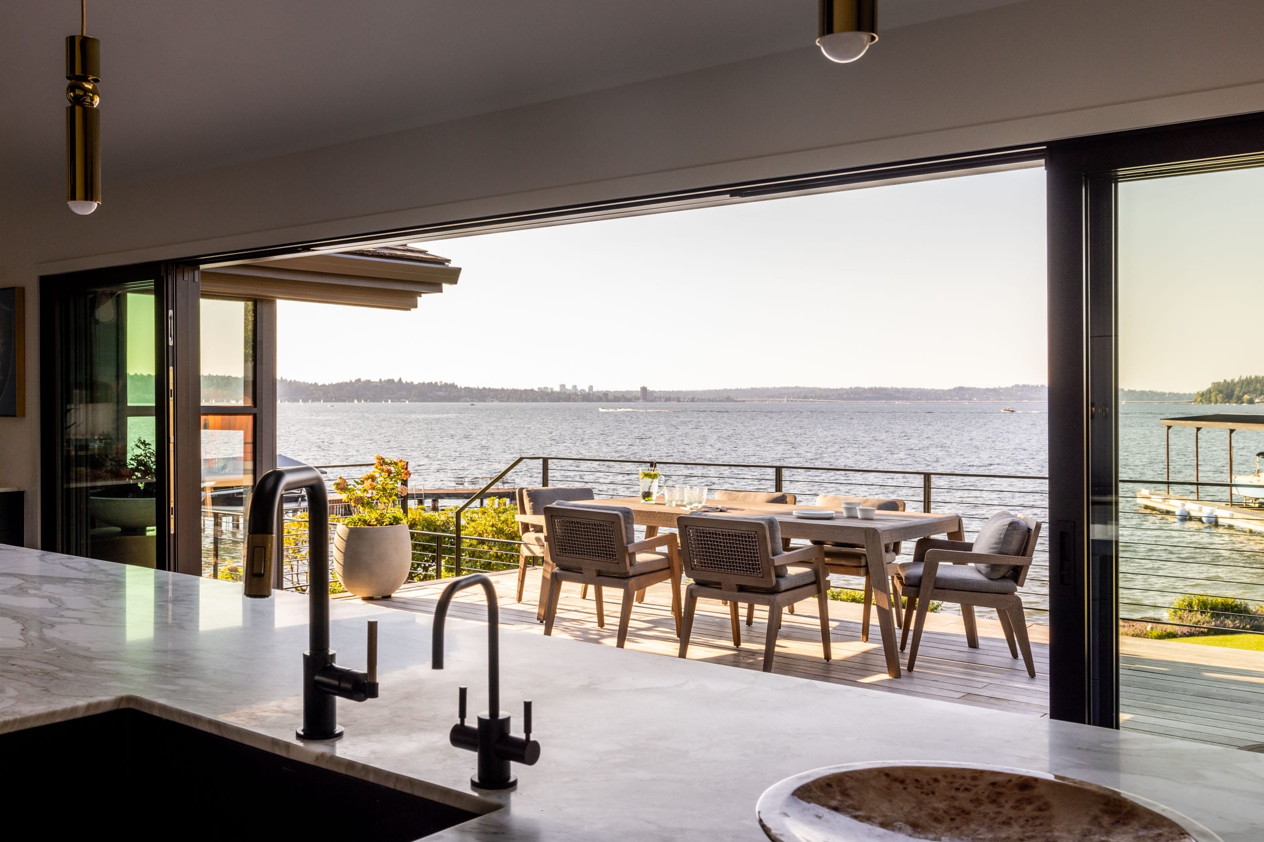 Modern kitchen with a view of a spacious patio featuring a dining set overlooking a large body of water and distant shoreline.