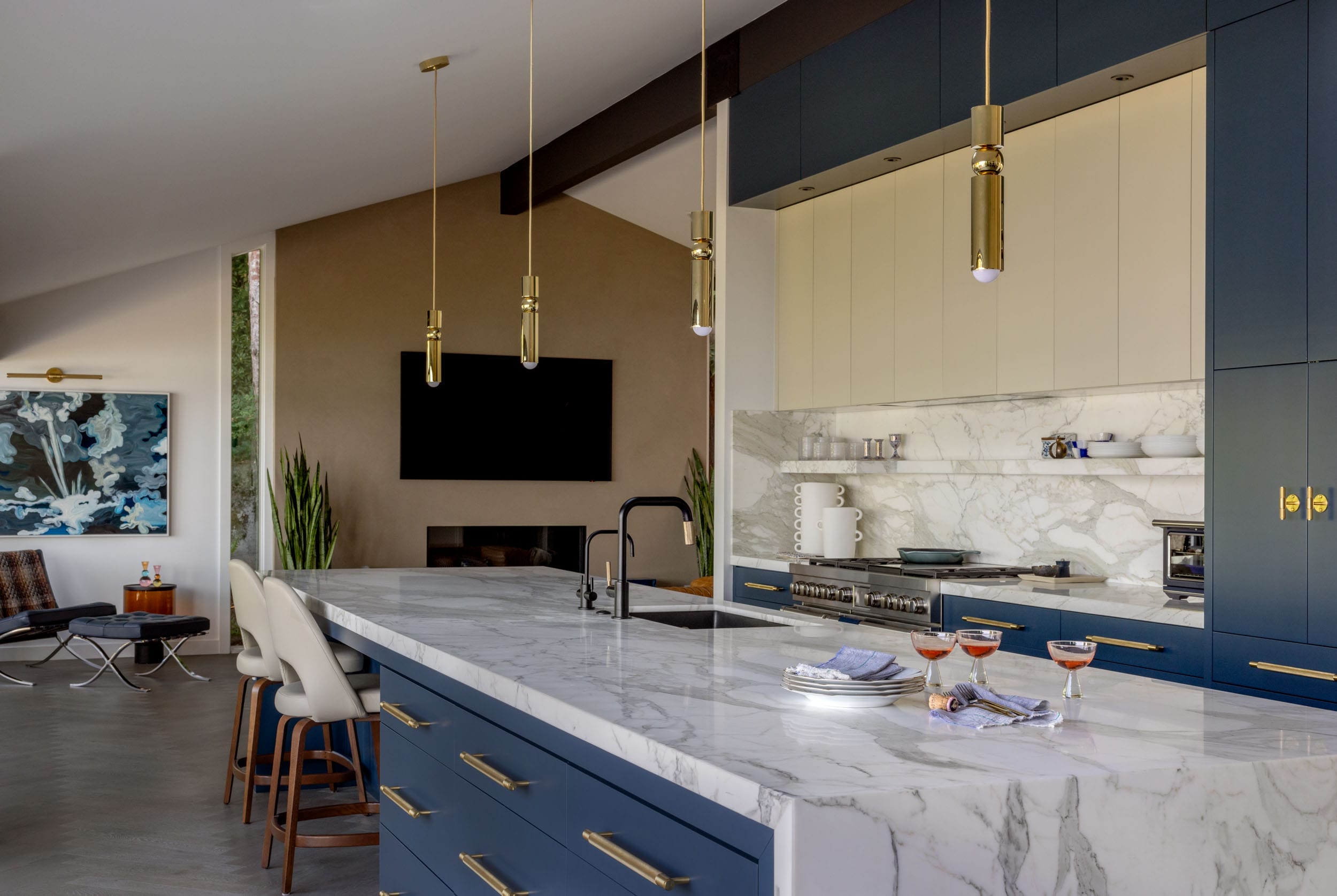 Modern kitchen with a marble island, blue cabinets, gold handles, and pendant lights. Stools line the island. A living area with a TV and artwork is visible in the background.