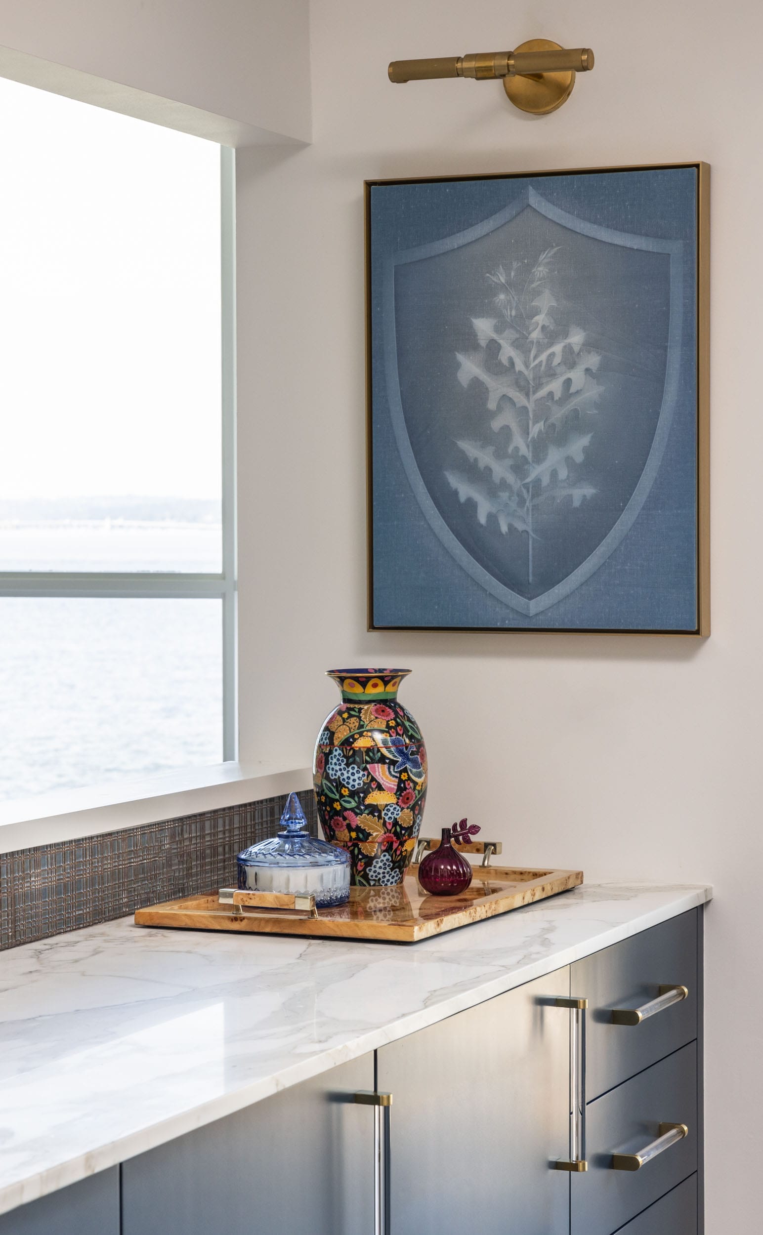 A modern kitchen corner features a marble countertop with decorative items, including a colorful vase. A framed blue artwork with a leaf design hangs on the wall above.