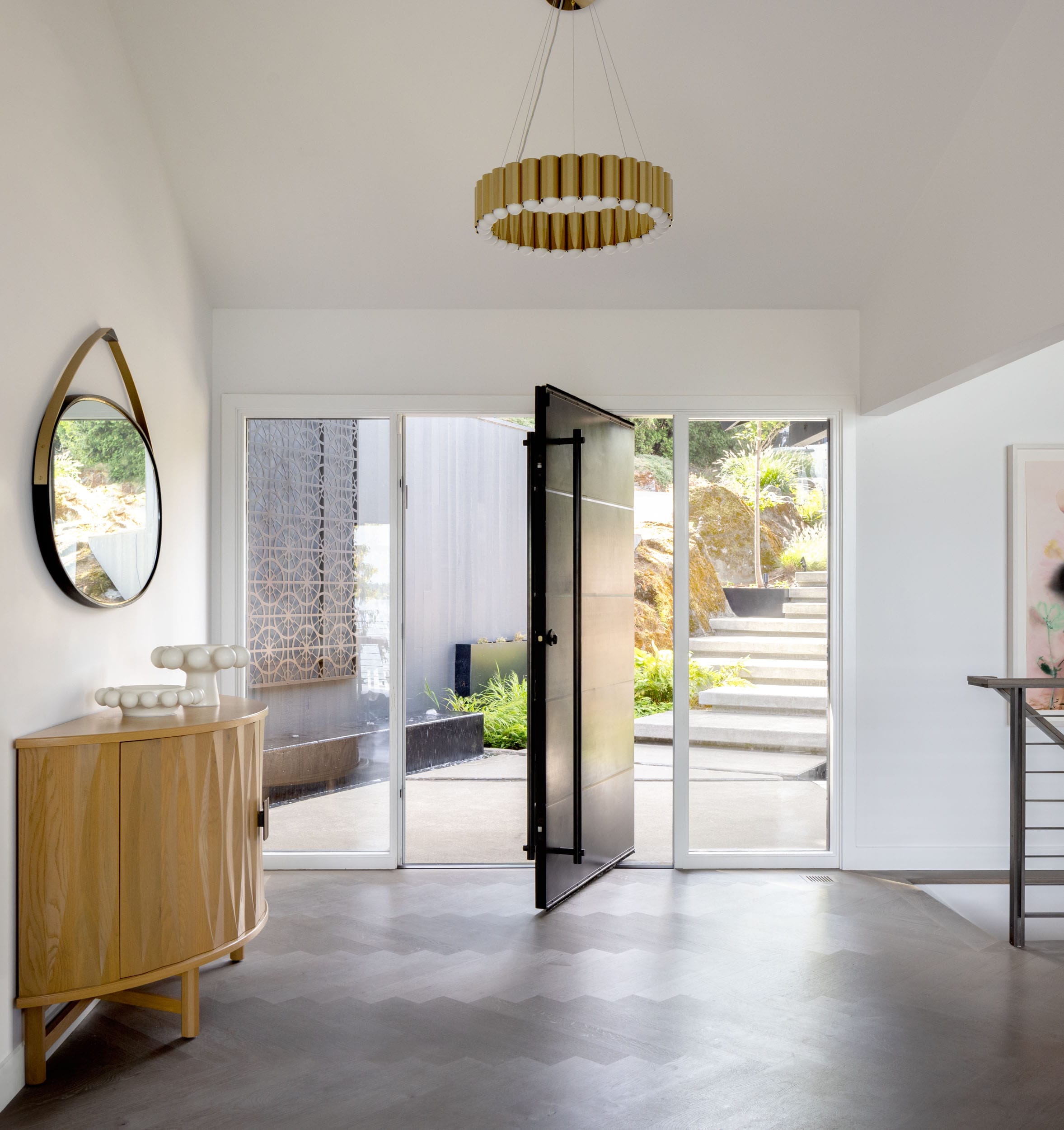 Modern entryway with an open black door, a round mirror, a wooden cabinet, and a chandelier. Outside, there are steps leading up with greenery visible through the glass.