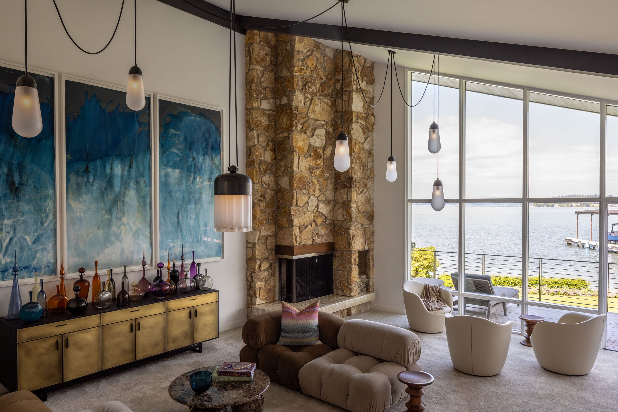 A modern living room with large windows, stone fireplace, plush seating, pendant lights, and blue abstract art on the wall. The view outside features a body of water and a pier.