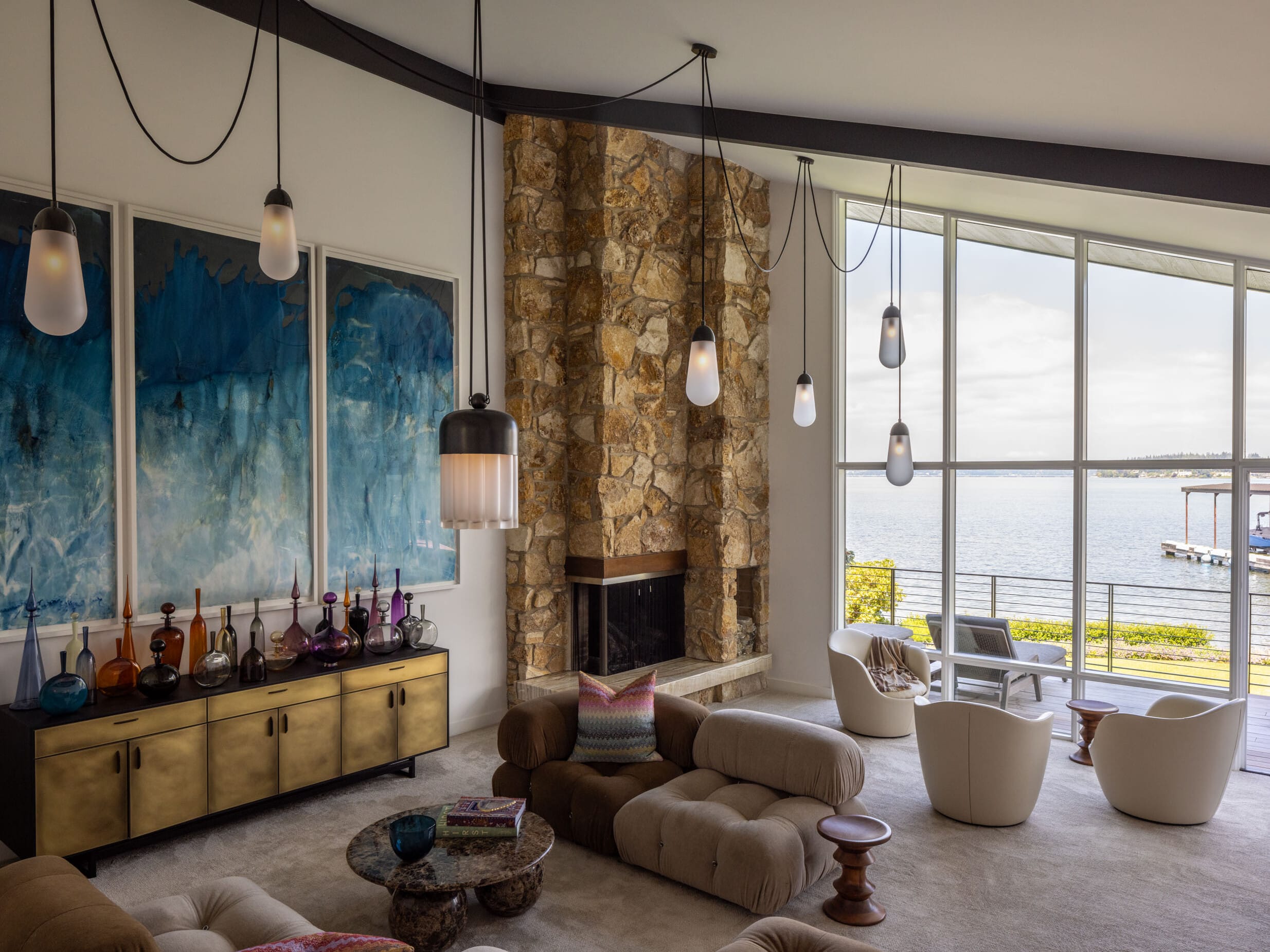 A modern living room with large windows, stone fireplace, plush seating, pendant lights, and blue abstract art on the wall. The view outside features a body of water and a pier.