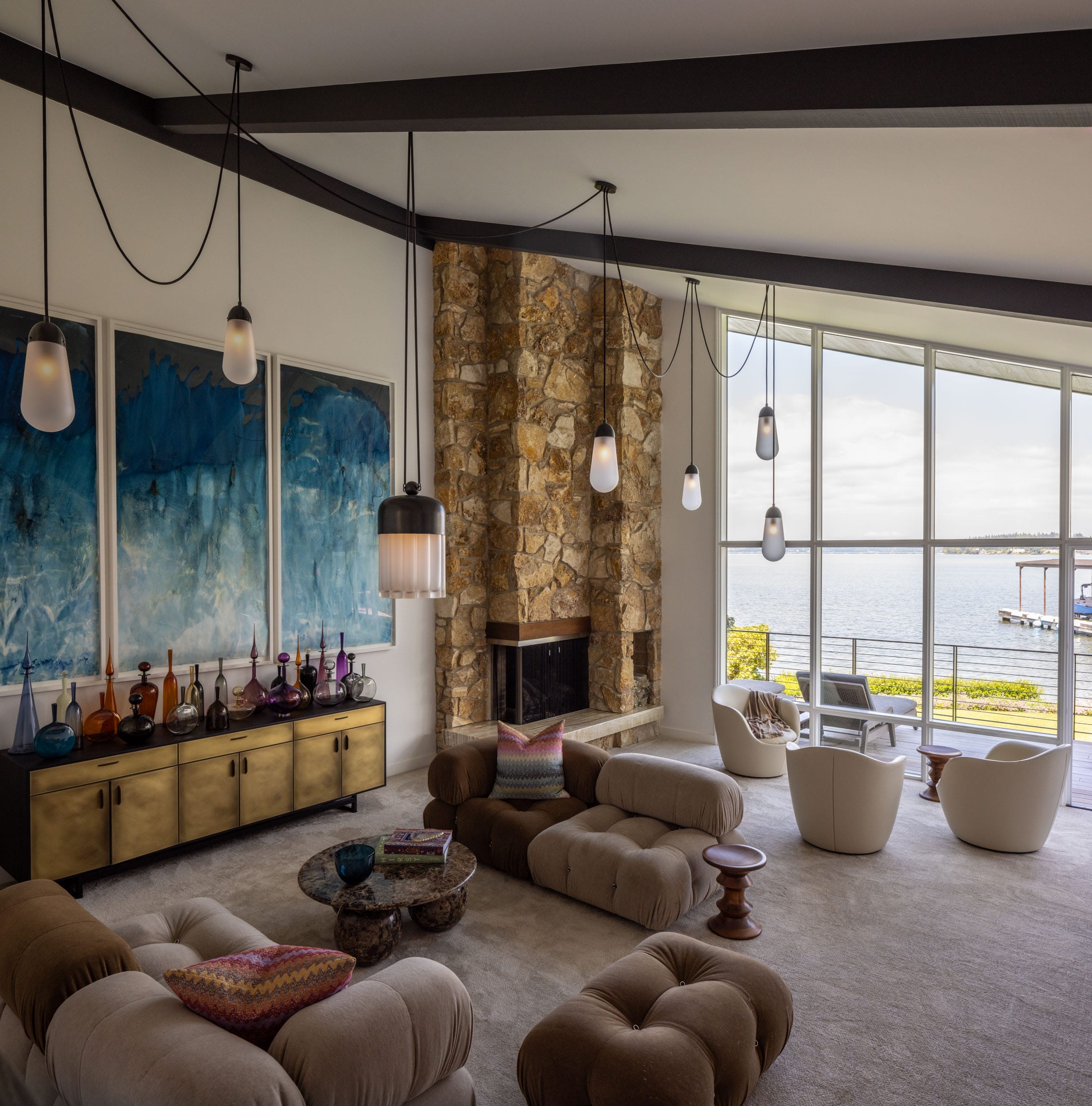 A modern living room with large windows, stone fireplace, plush seating, pendant lights, and blue abstract art on the wall. The view outside features a body of water and a pier.