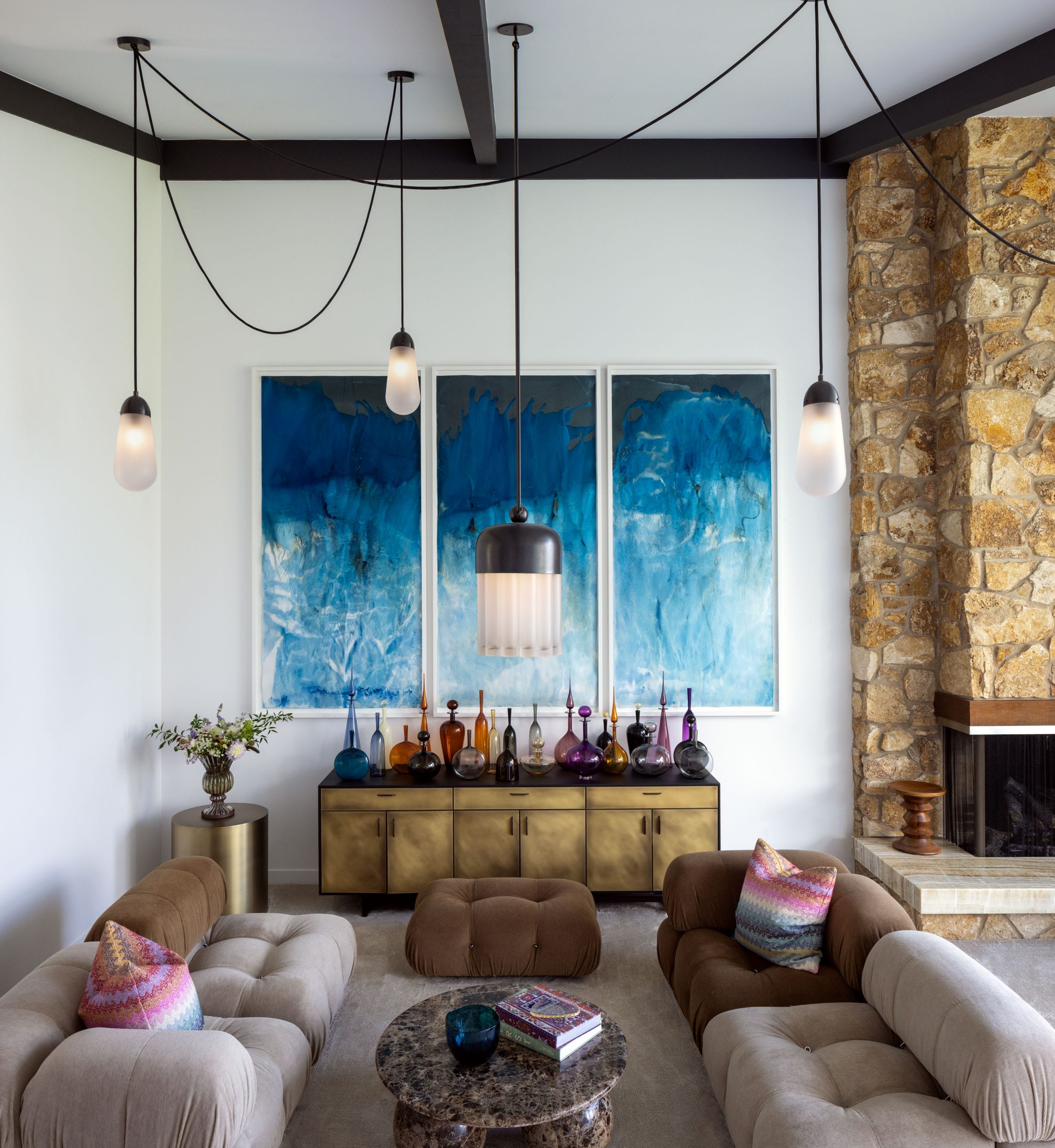 Modern living room with beige sofas, a marble coffee table, and a stone fireplace. Blue abstract triptych art hangs above a console with decorative glass bottles. Hanging lights are overhead.