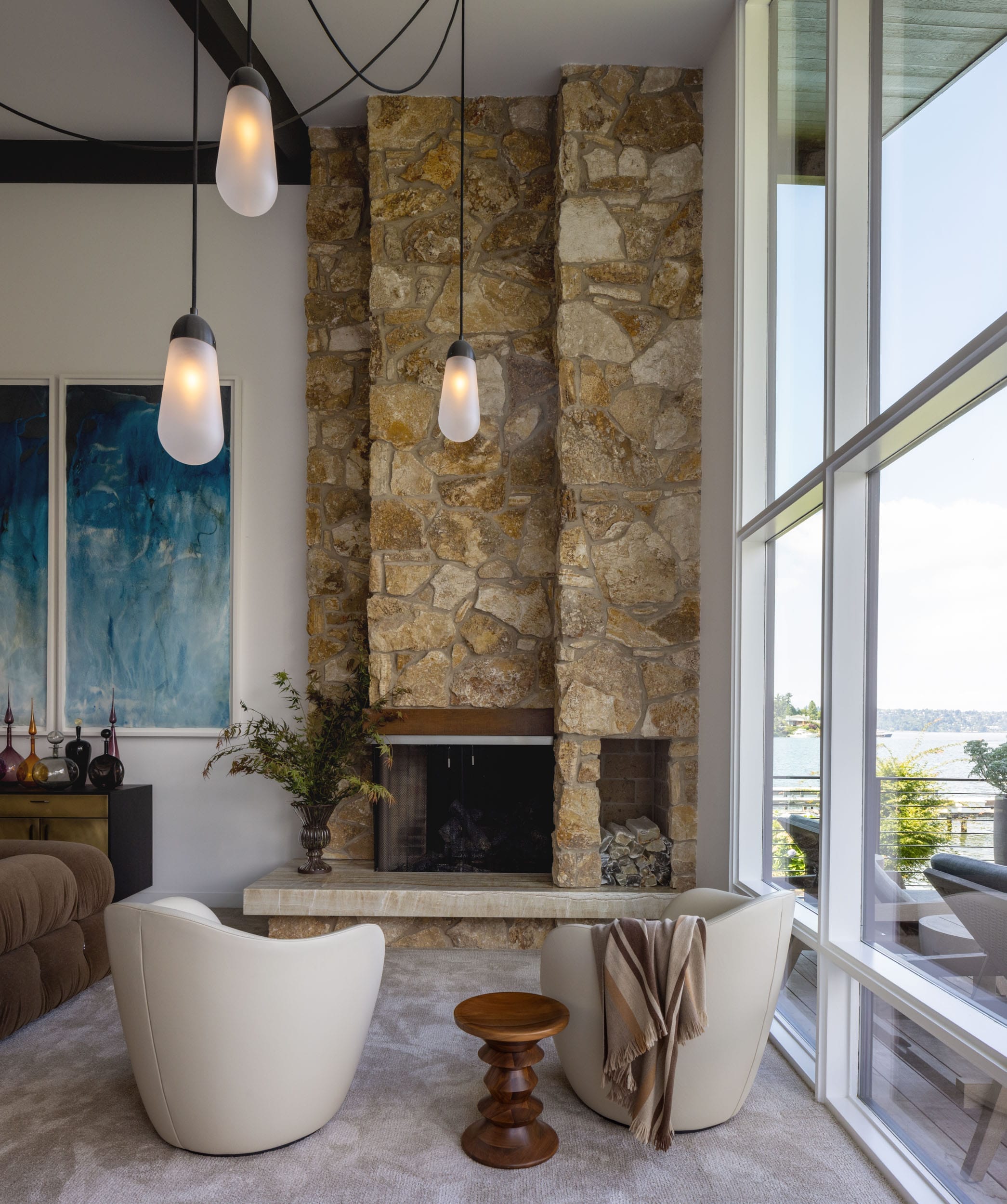 A modern living room with a stone fireplace, two white chairs, a brown wooden table, and floor-to-ceiling windows overlooking a water view.