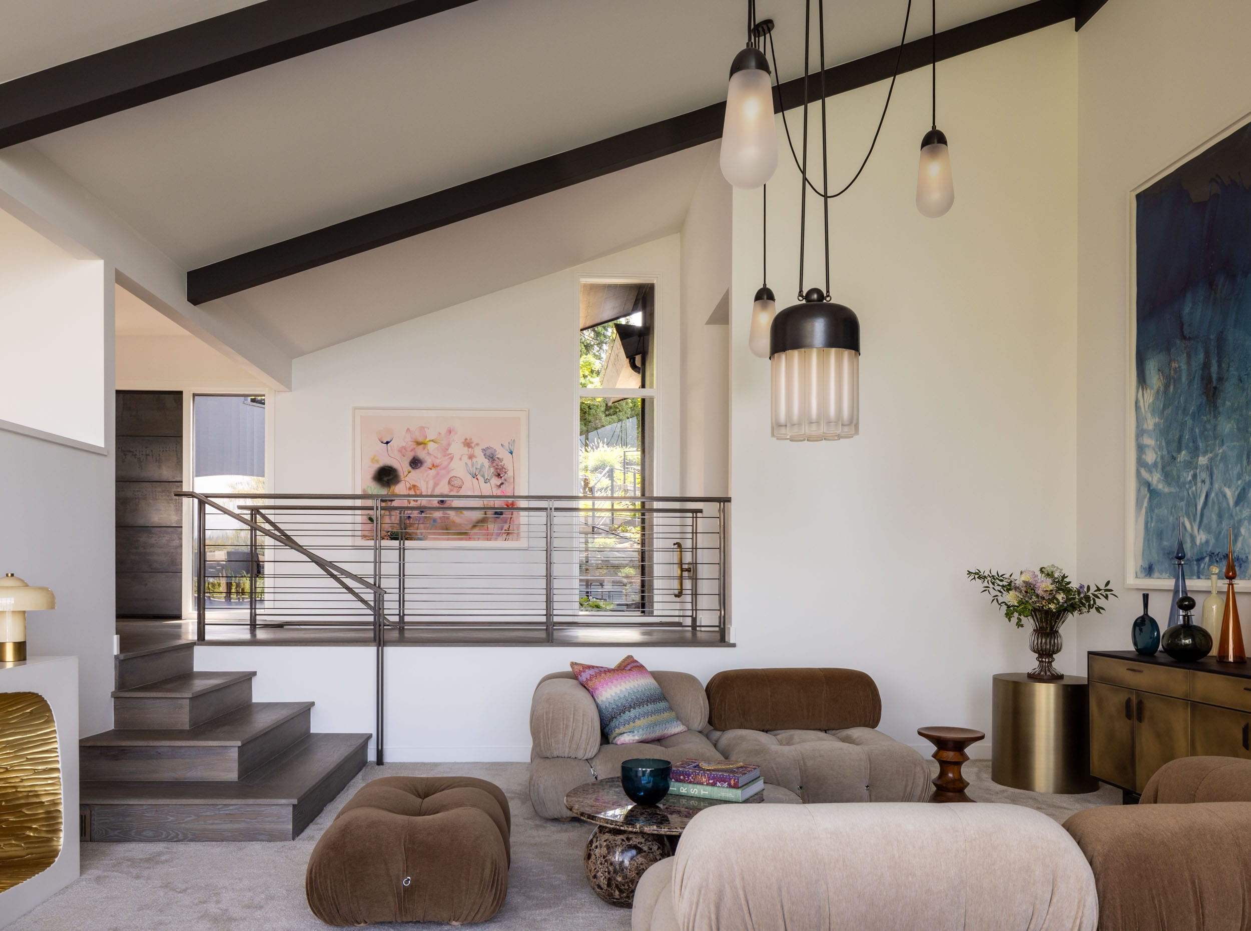 A modern living room with brown seating, a low table, pendant lights, artwork, and a metallic railing by the stairs.