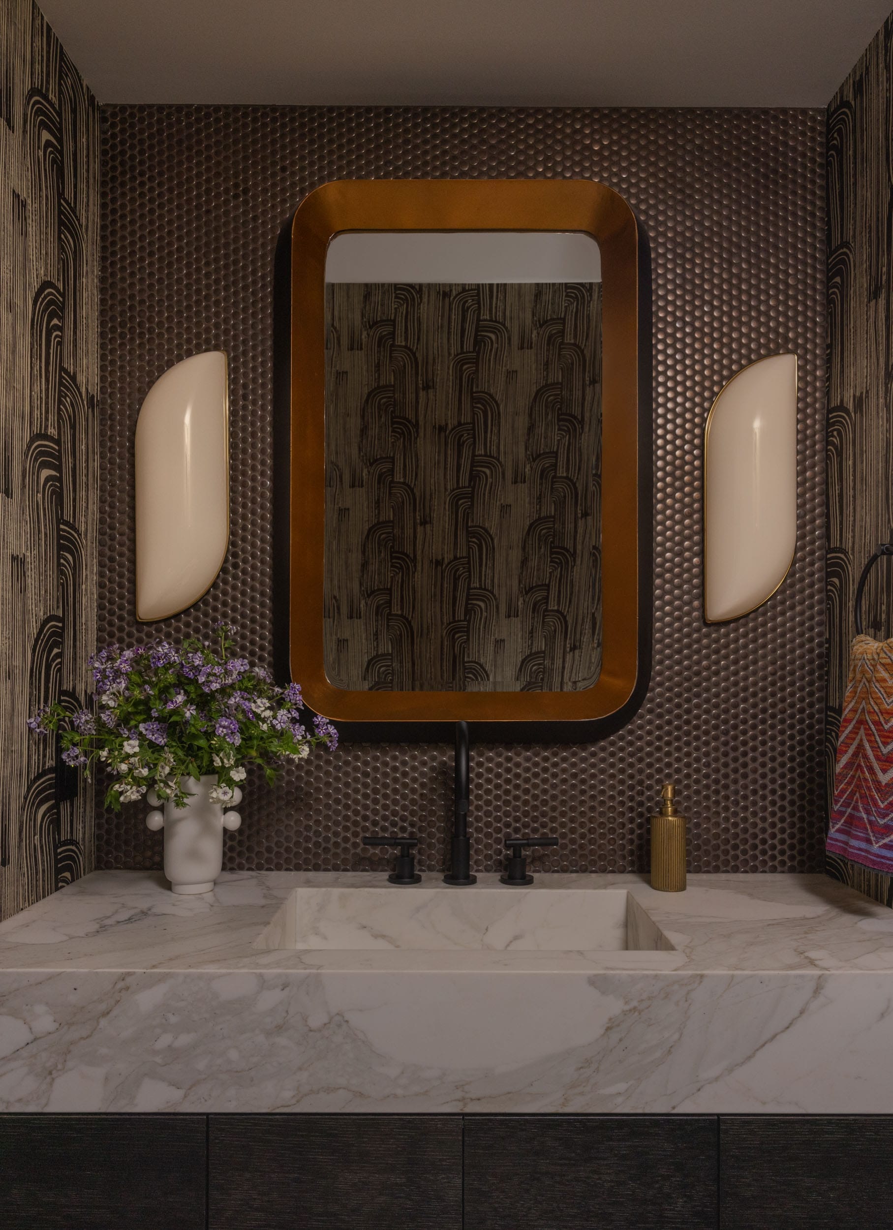 A modern bathroom with a marble sink, a large rectangular mirror, black faucets, a vase of purple flowers, and decorative wallpaper.