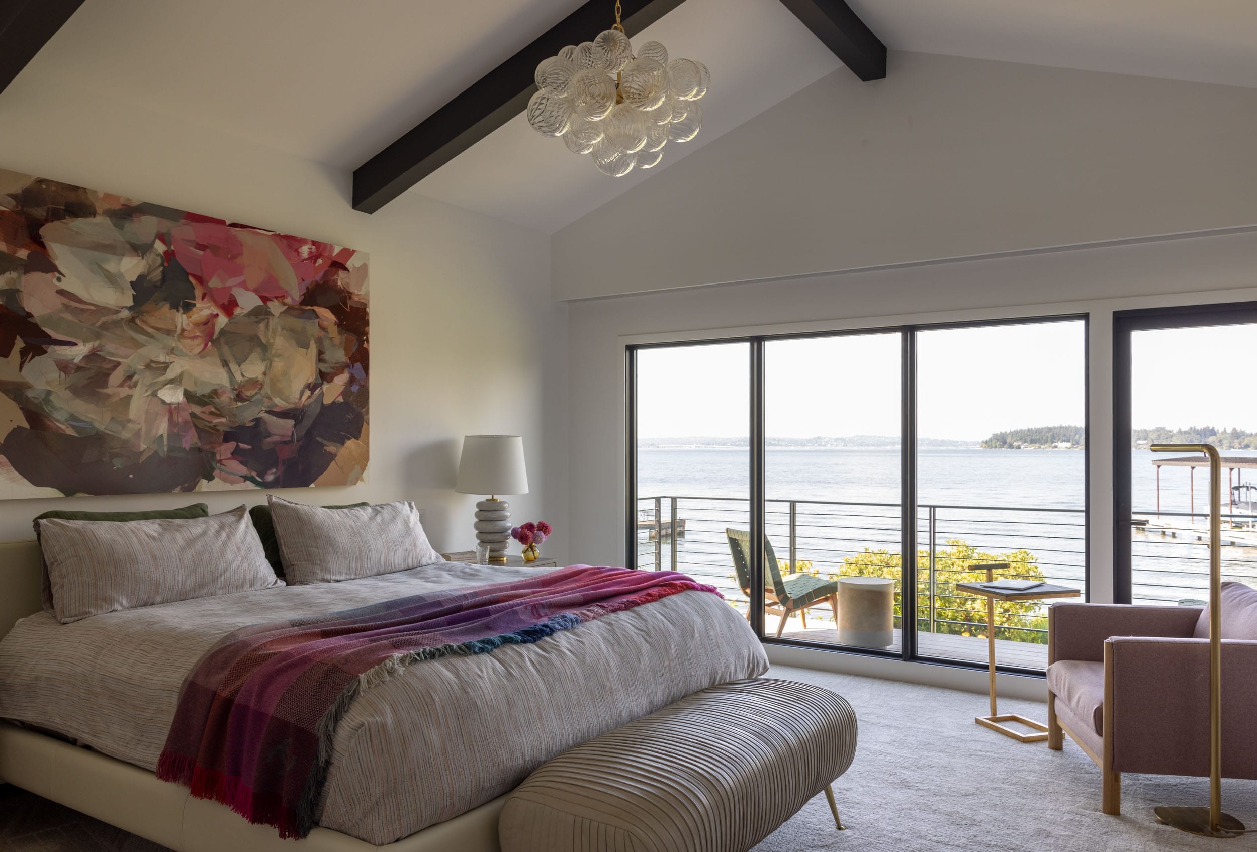 Bedroom with large window overlooking a lake, featuring a bed with a colorful blanket, abstract wall art, an upholstered bench, pink chair, and a modern glass chandelier.