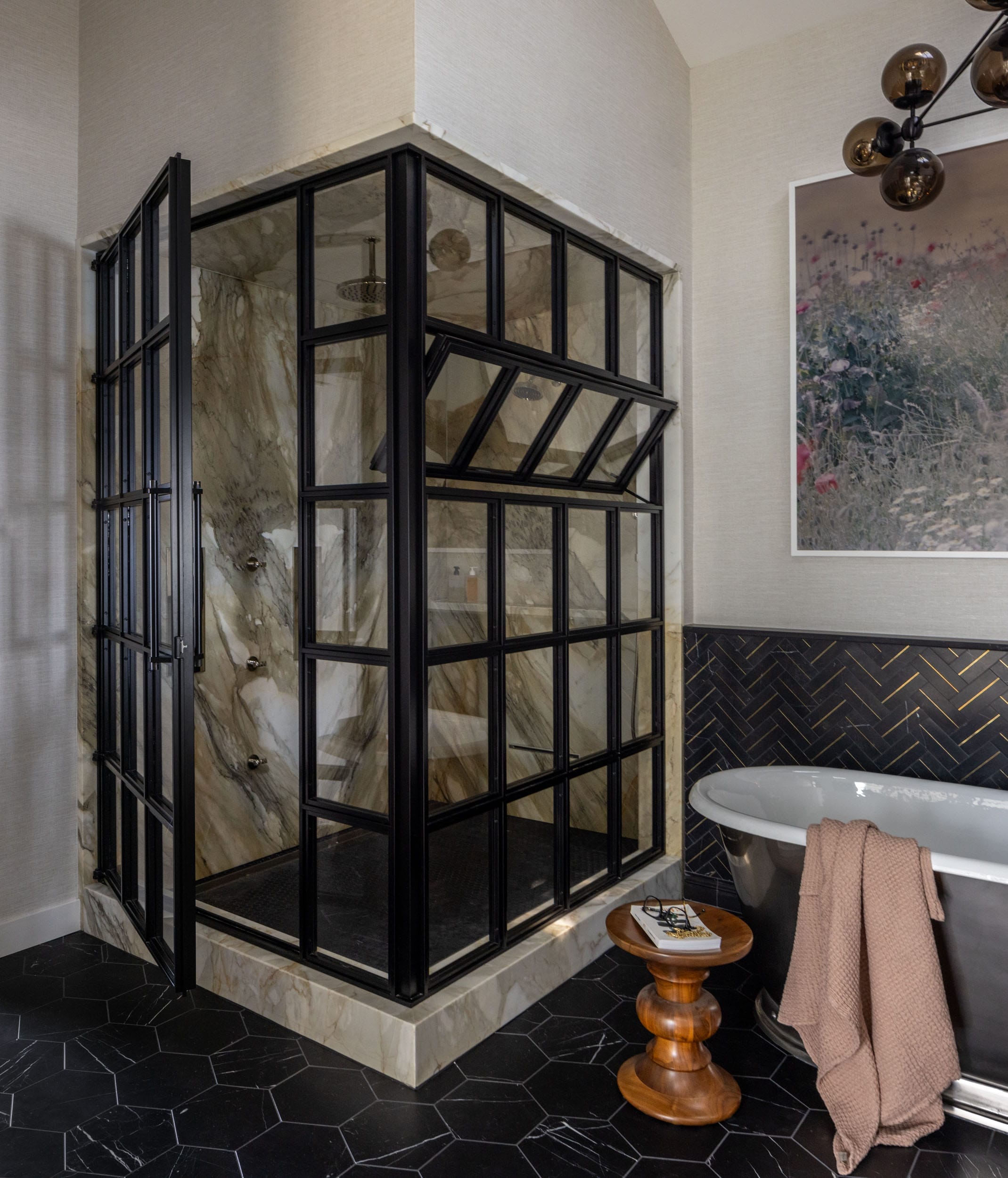 Modern bathroom with a glass-enclosed shower, freestanding bathtub, and geometric black floor tiles. A wooden side table holds a folded brown towel. Wall art and light fixture are visible.