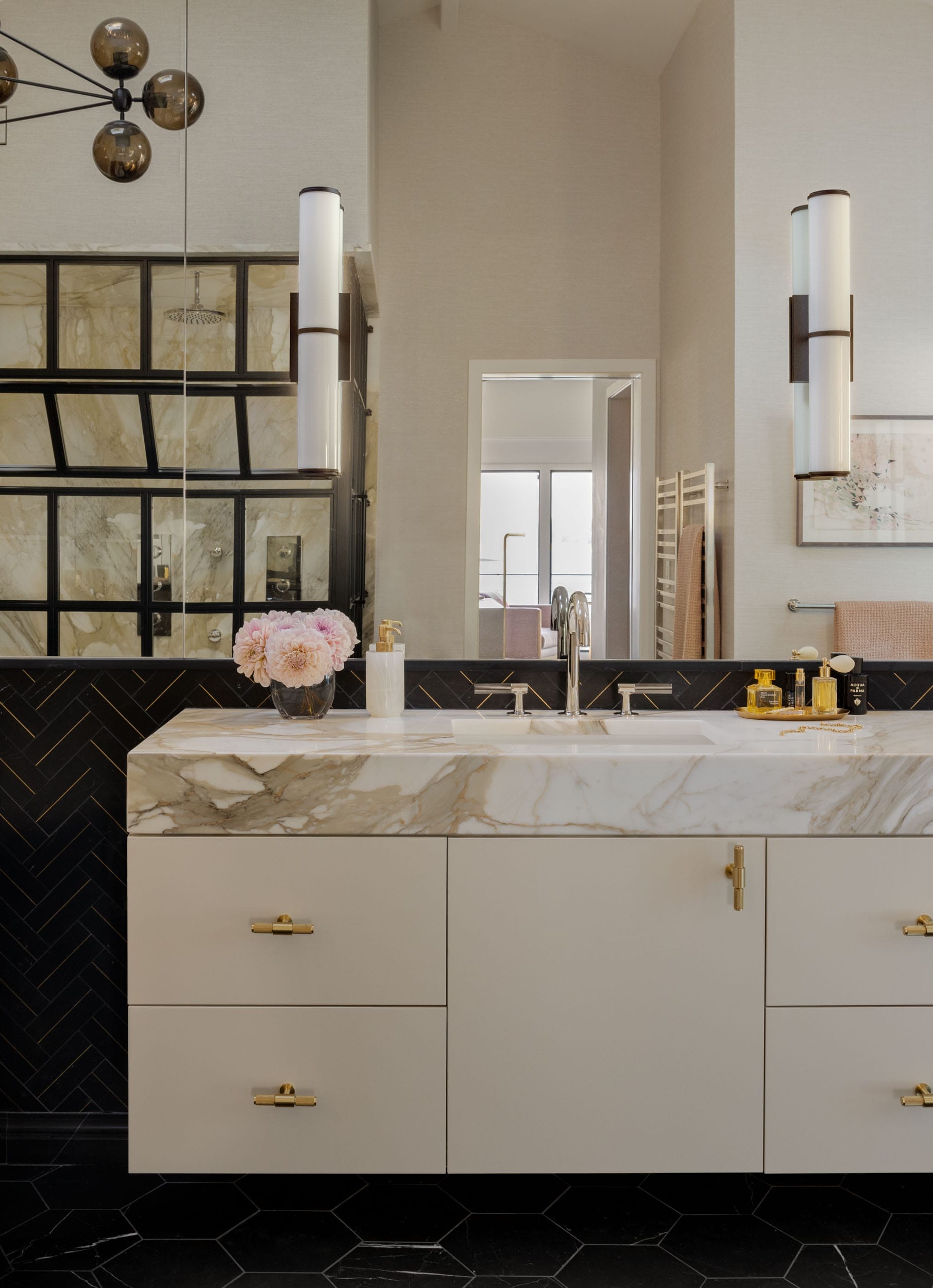 Modern bathroom with a marble-topped vanity, gold fixtures, black herringbone tiles, double sink, and a glass shower door. Pink flowers in a vase add a touch of color.