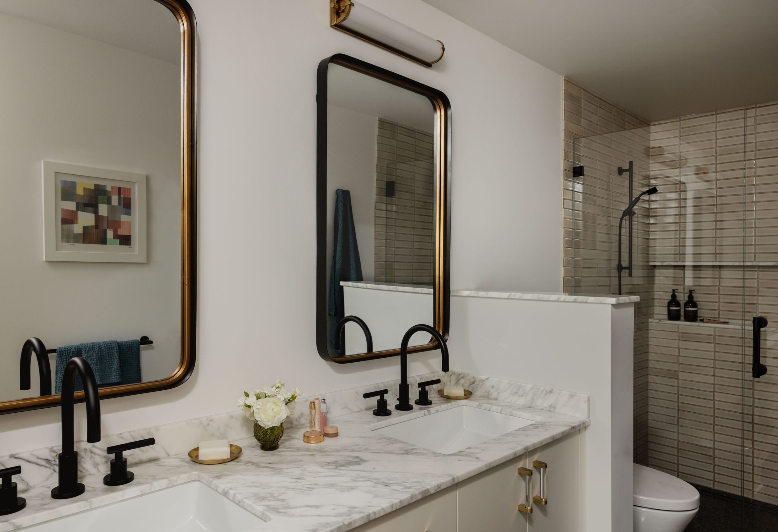 Bathroom with two sinks, marble countertop, black faucets, and two rectangular mirrors. Glass shower enclosure with beige tiles in the background and a small wall art piece on the left.