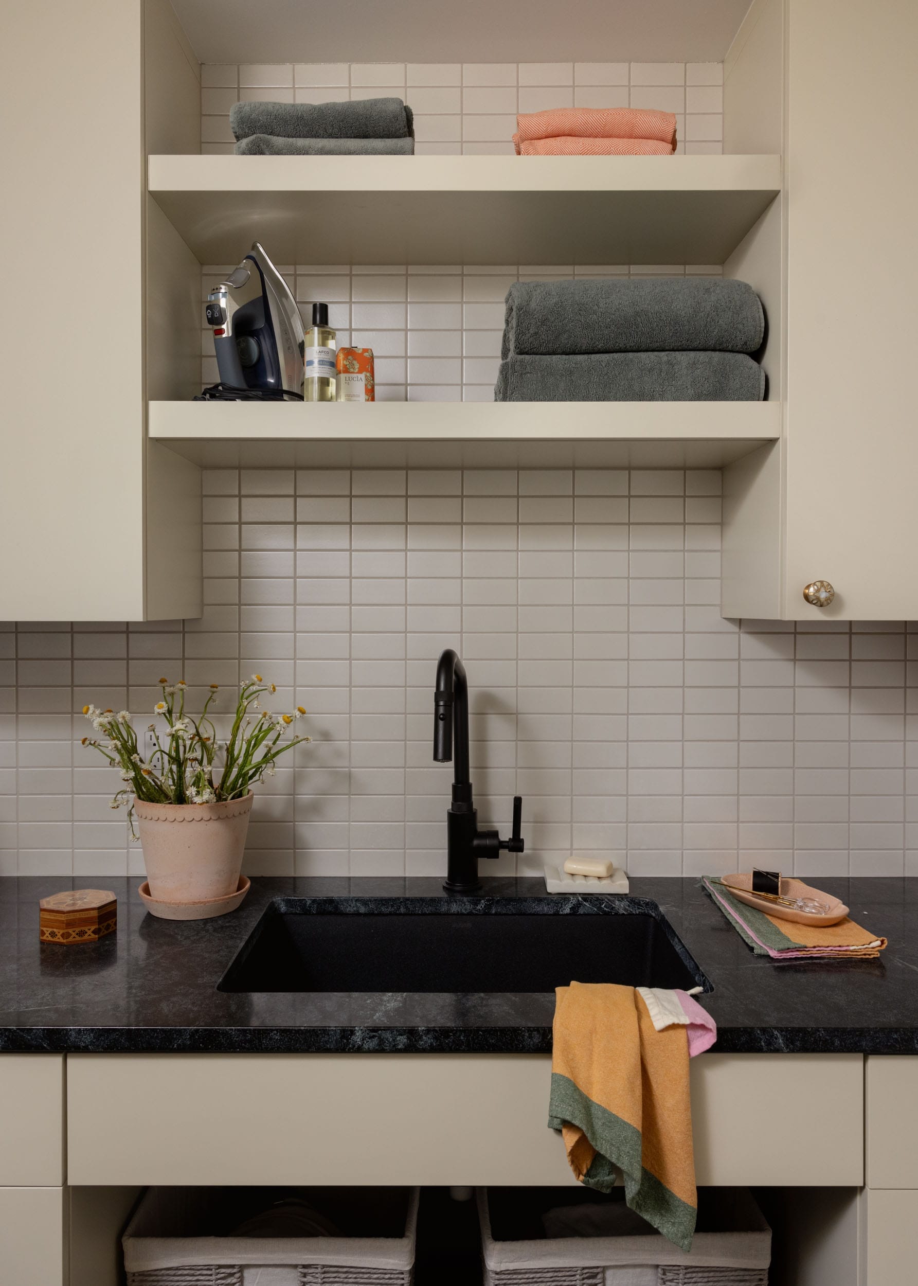 A beige laundry room with a black sink and faucet. Shelves hold towels, an iron, and cleaning supplies. A potted plant and colorful cloth are near the sink.