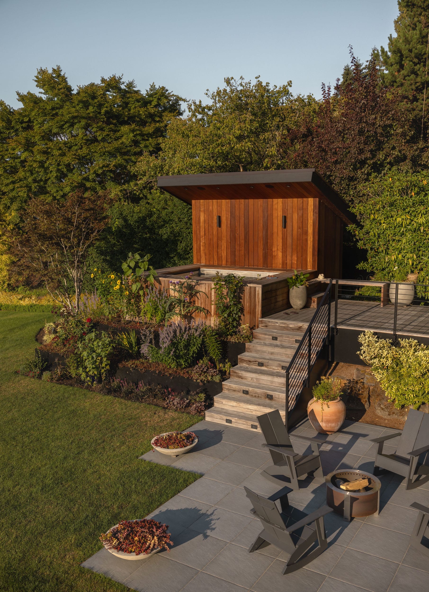 A modern backyard with a wooden shed, elevated deck, stairs, and patio. Surrounded by trees and plants, with two chairs and potted plants on the patio.