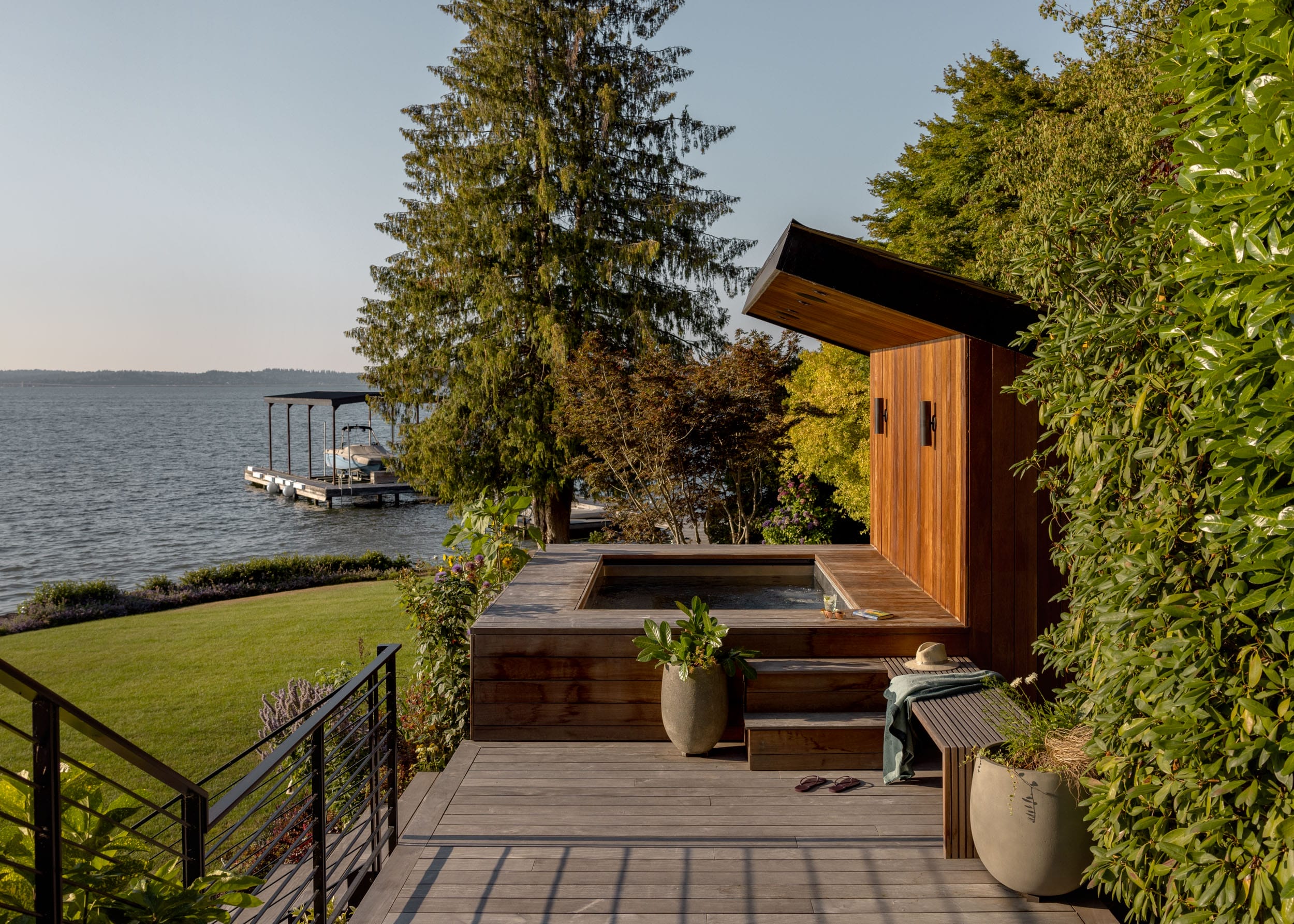 Wooden deck with hot tub overlooking a lake, surrounded by lush greenery. Steps lead down to a grassy lawn and a dock with a covered boat area.