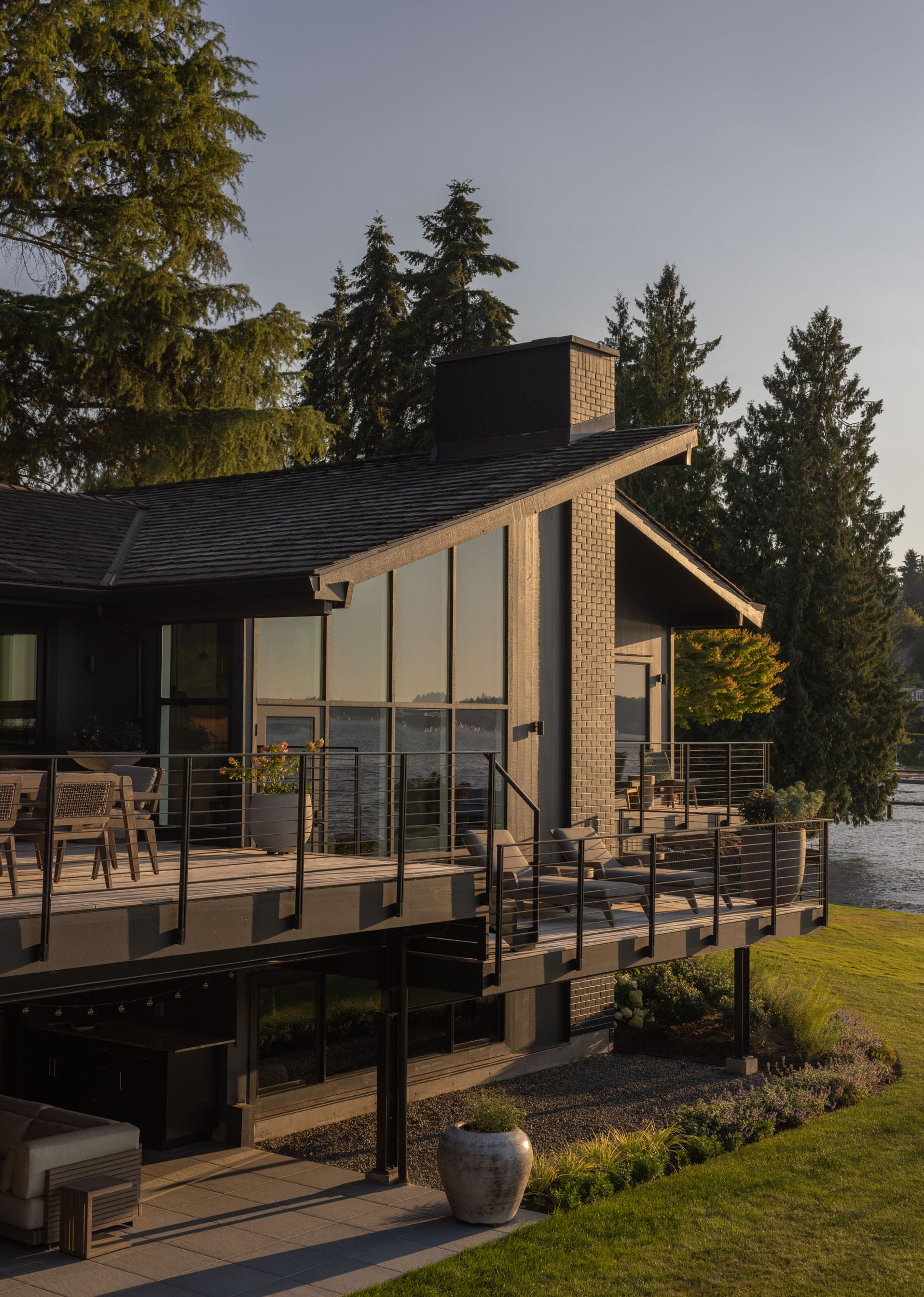 Modern lakeside house with large windows, a spacious deck with outdoor furniture, and surrounded by trees. A body of water is visible in the background.