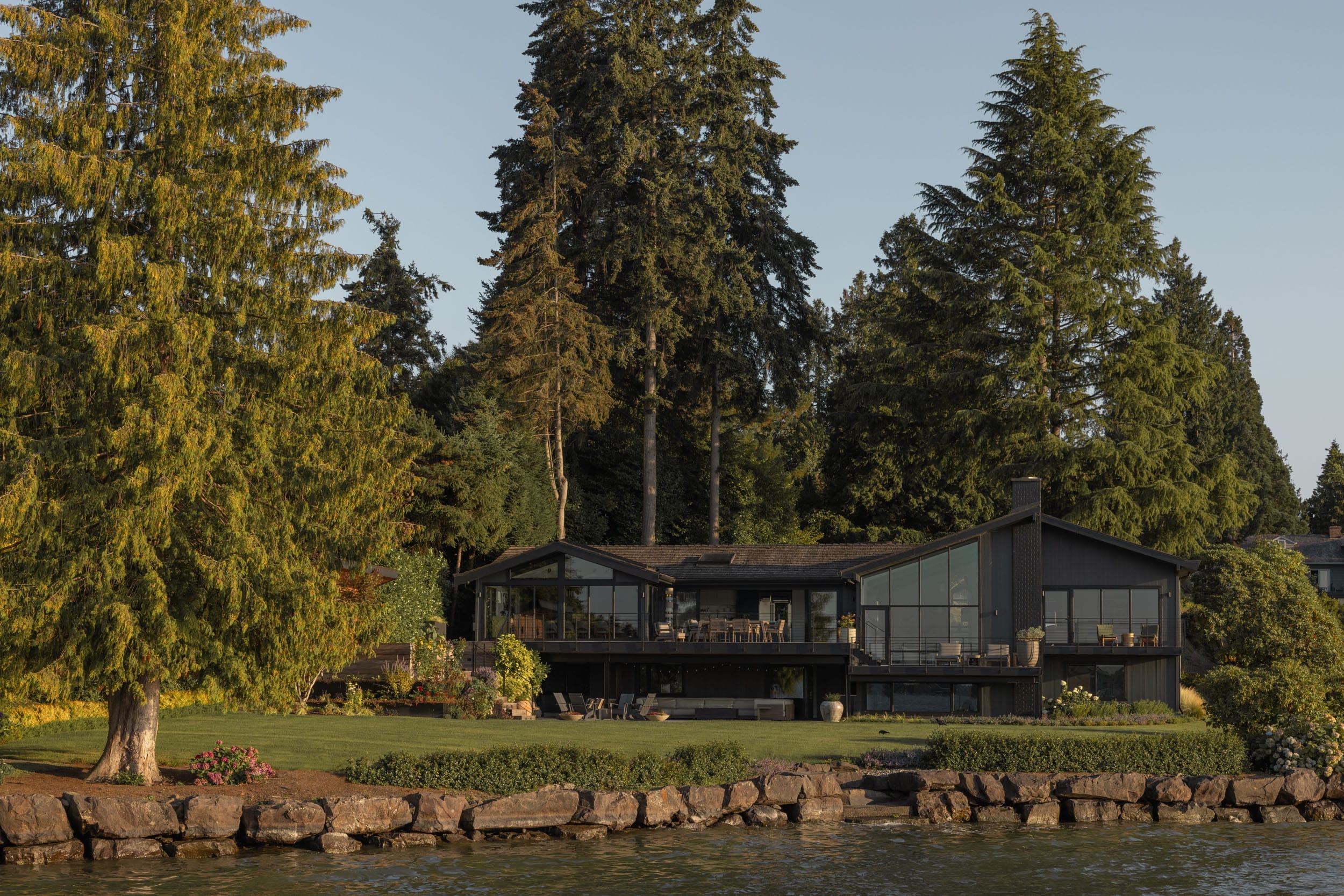 A modern black house surrounded by tall trees, with a spacious lawn, sits by a rocky shoreline under a clear sky.