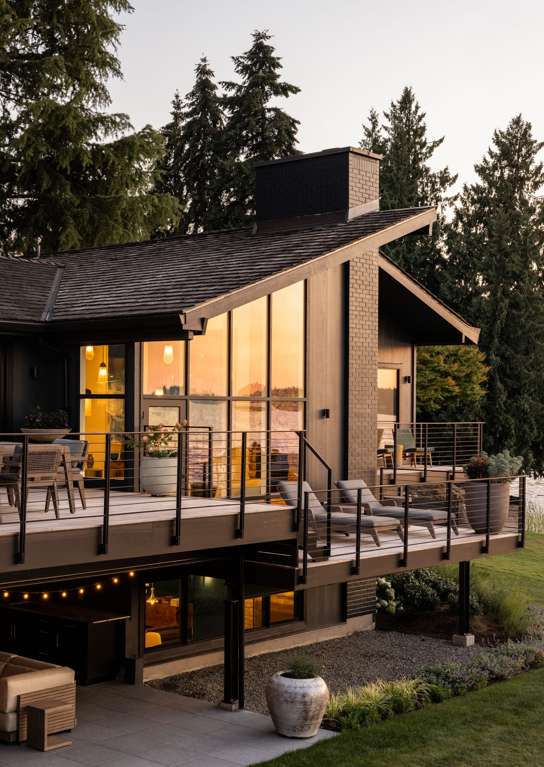 Modern house with large windows and a wooden deck, surrounded by trees. A patio with furniture is visible on the deck, and the sunset reflects on the glass.