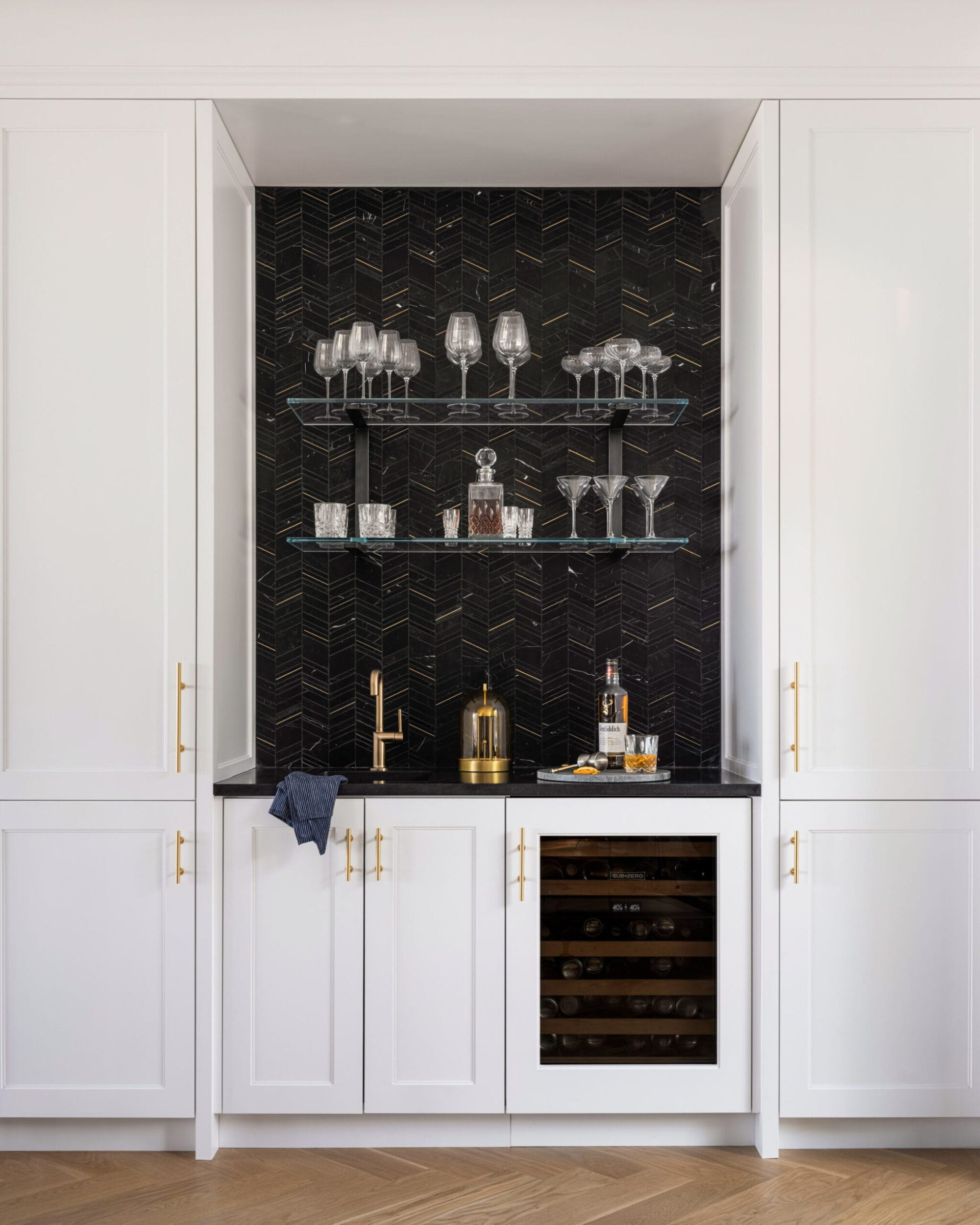 A small home bar with white cabinets, a black chevron backsplash, glass shelves with glasses, a drink shaker, and a wine cooler.