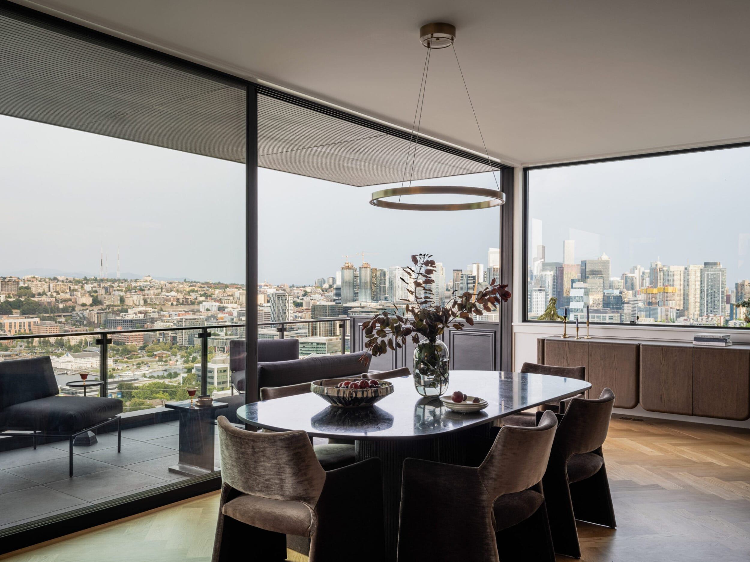 A modern dining room with a large table, eight chairs, and a vase of flowers, overlooking a city skyline through floor-to-ceiling windows.