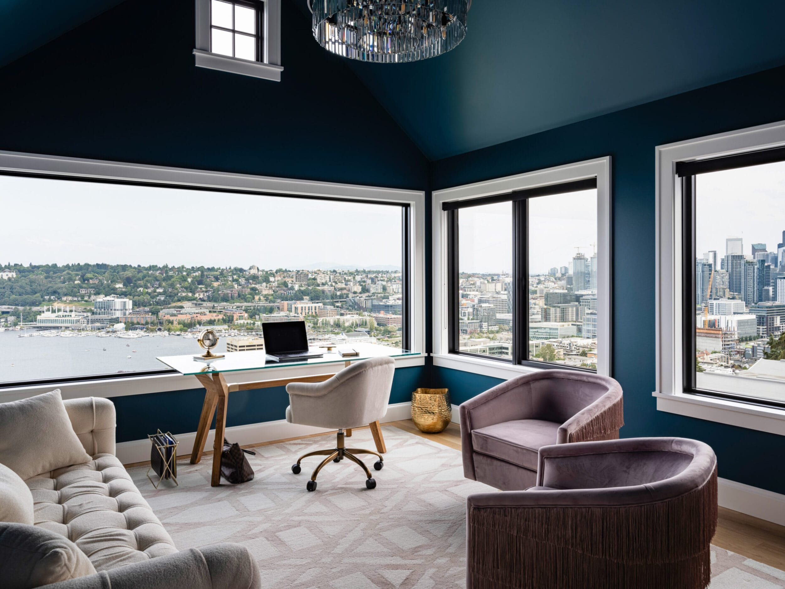 A modern office with large windows showing a cityscape and water view, featuring a desk, chair, sofa, and armchair, in a room with dark blue walls and a chandelier.