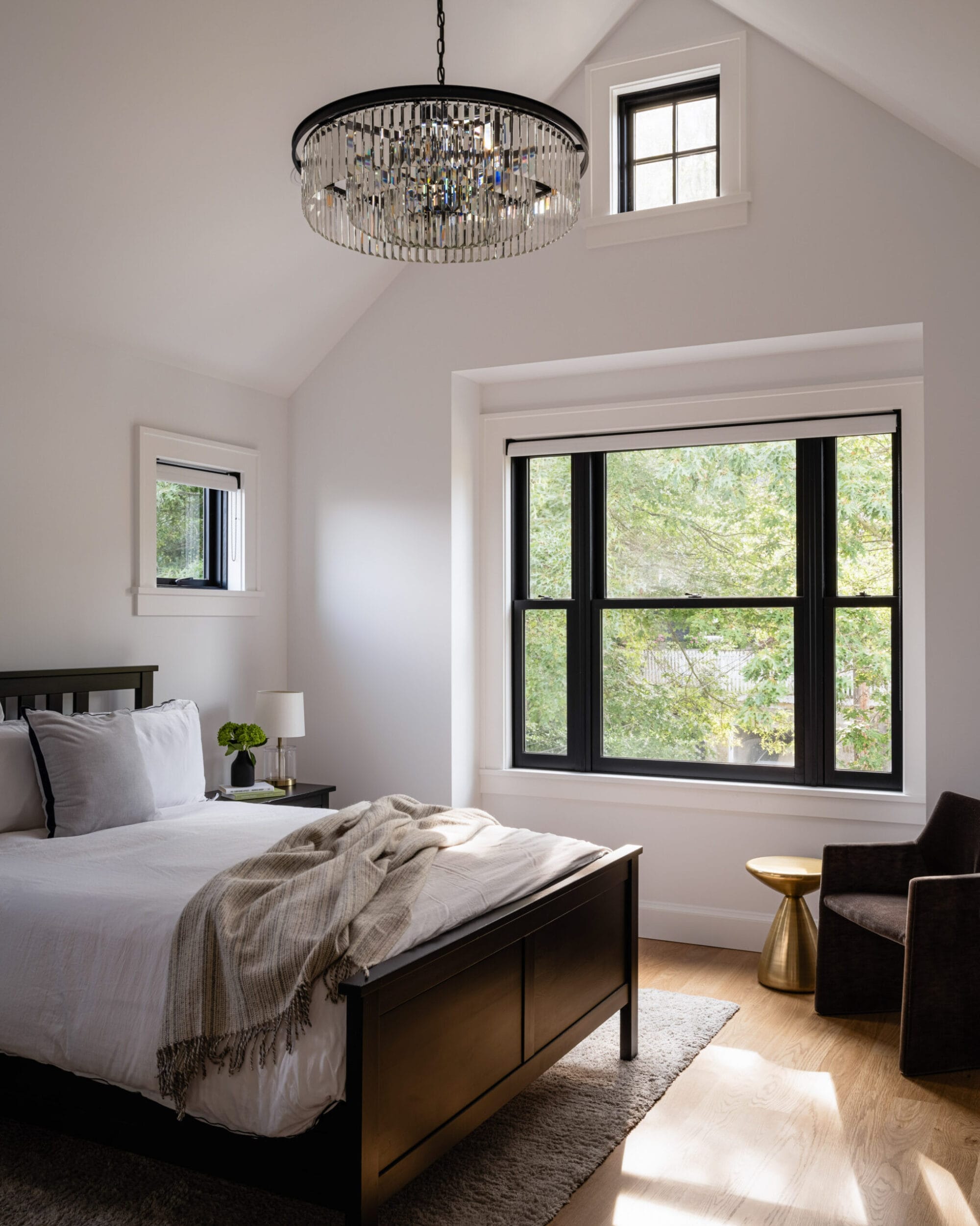 Bedroom with a vaulted ceiling, large windows, and a modern chandelier. Features a black bed, gray bedding, side tables, lamp, and a chair. Light wooden floor and view of greenery outside.