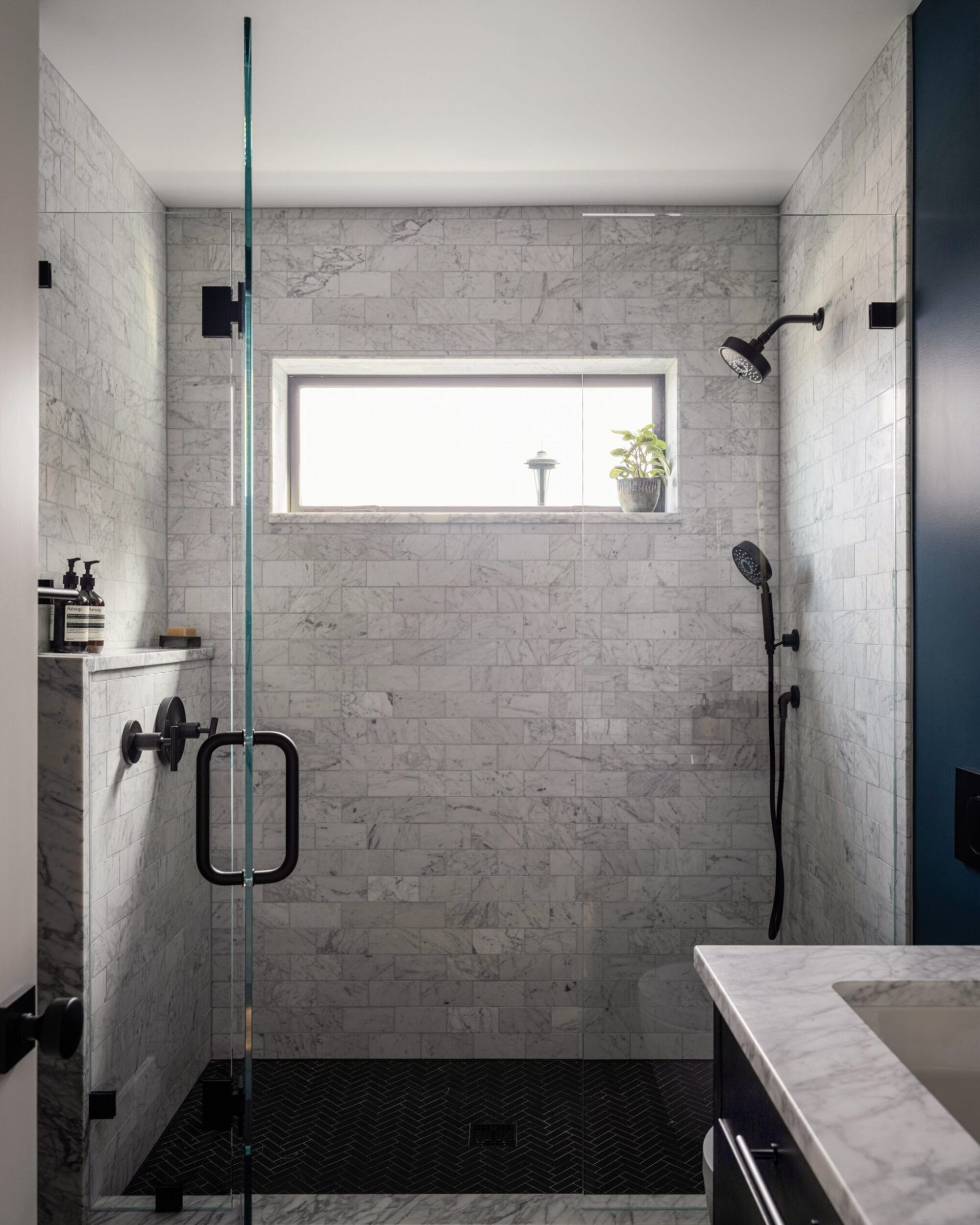 Modern bathroom shower with gray marble tiles, a glass door, and dual showerheads. A small window above allows natural light. A potted plant rests on the window ledge.