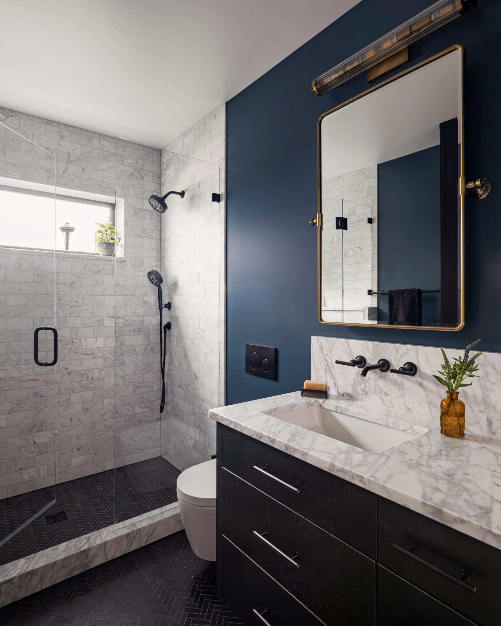 Modern bathroom with a glass-enclosed shower, marble tile walls, dark vanity with marble countertop, rectangular mirror, and dark blue wall. A small plant is placed on the countertop.