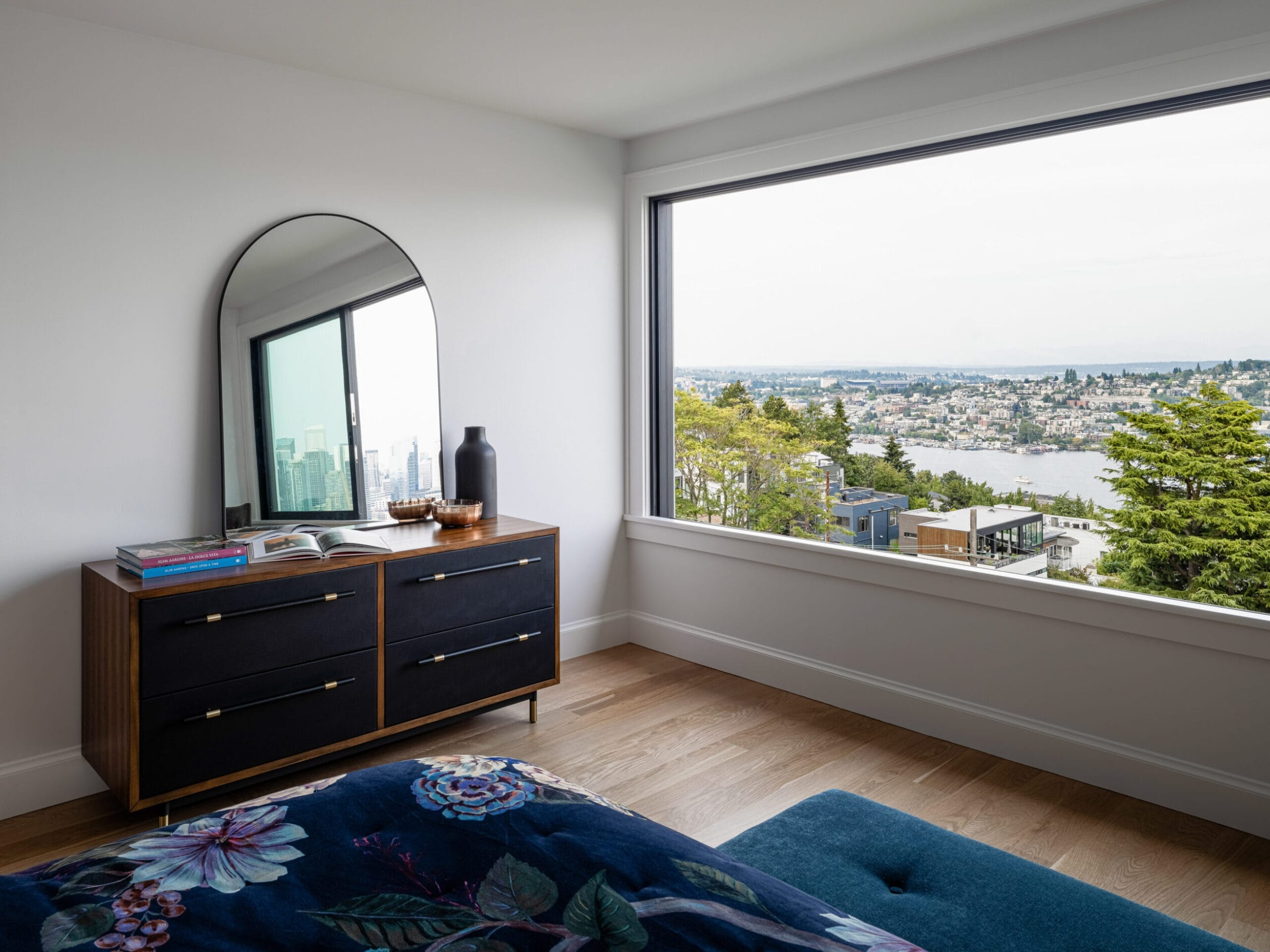 Modern bedroom with a wooden dresser, large round mirror, and a window offering a scenic view of trees and water.