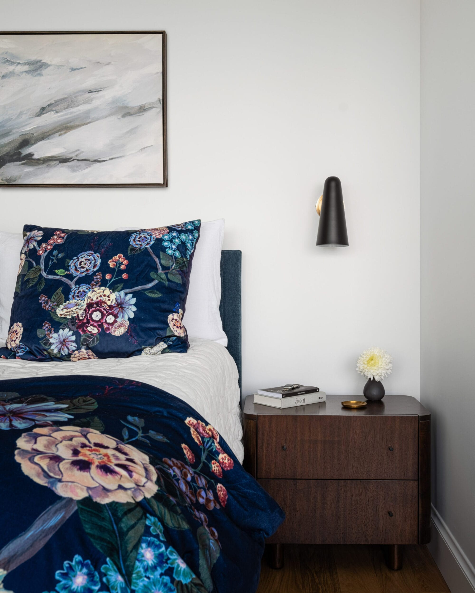 Bedroom with a floral-themed bedspread, wooden nightstand, books, a small lamp, and a flower in a vase. A wall lamp and abstract painting are above the bed.