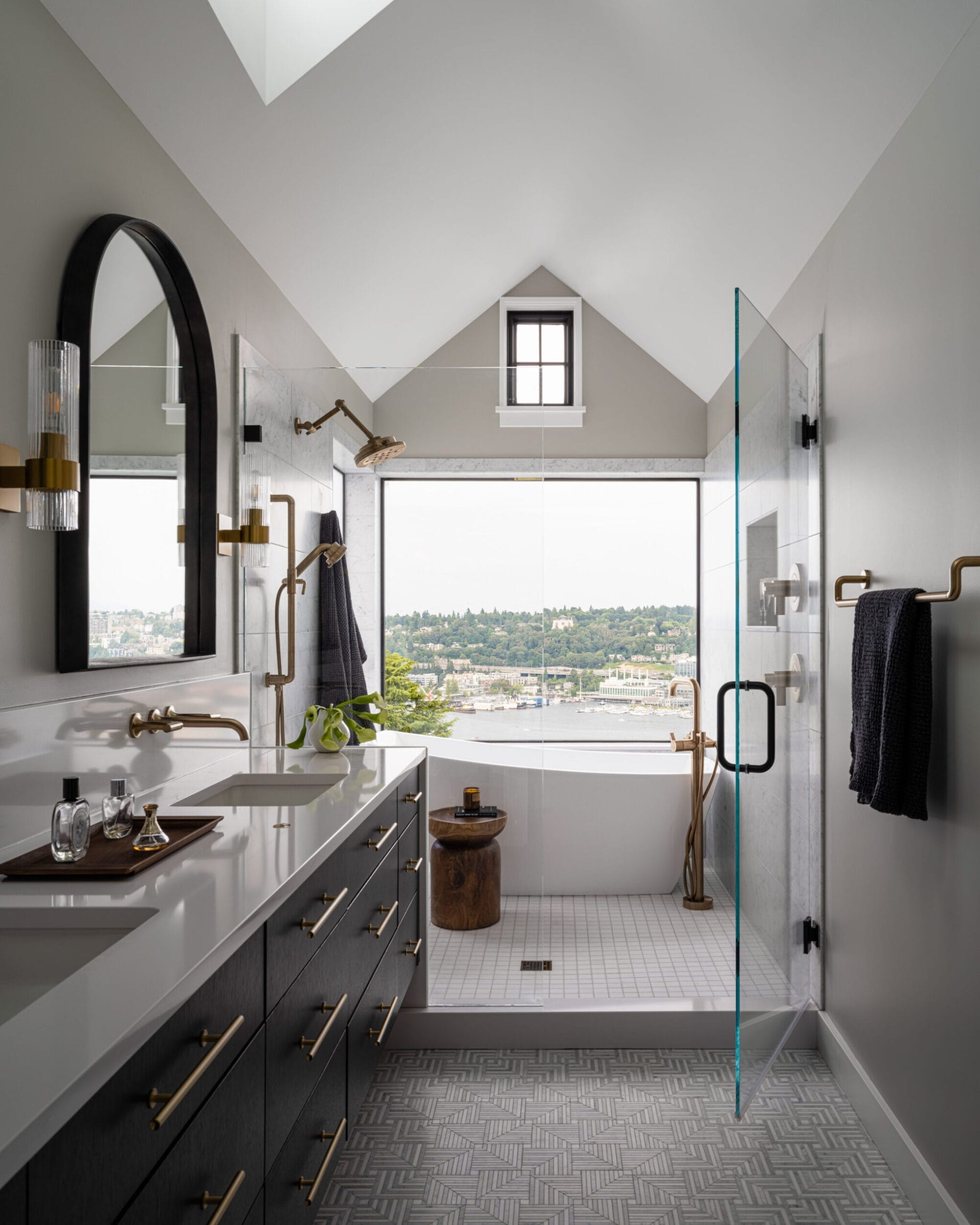 Modern bathroom with a large window, glass shower, freestanding tub, black vanity with dual sinks, and brass fixtures. Patterned floor tiles and skylight enhance the space.