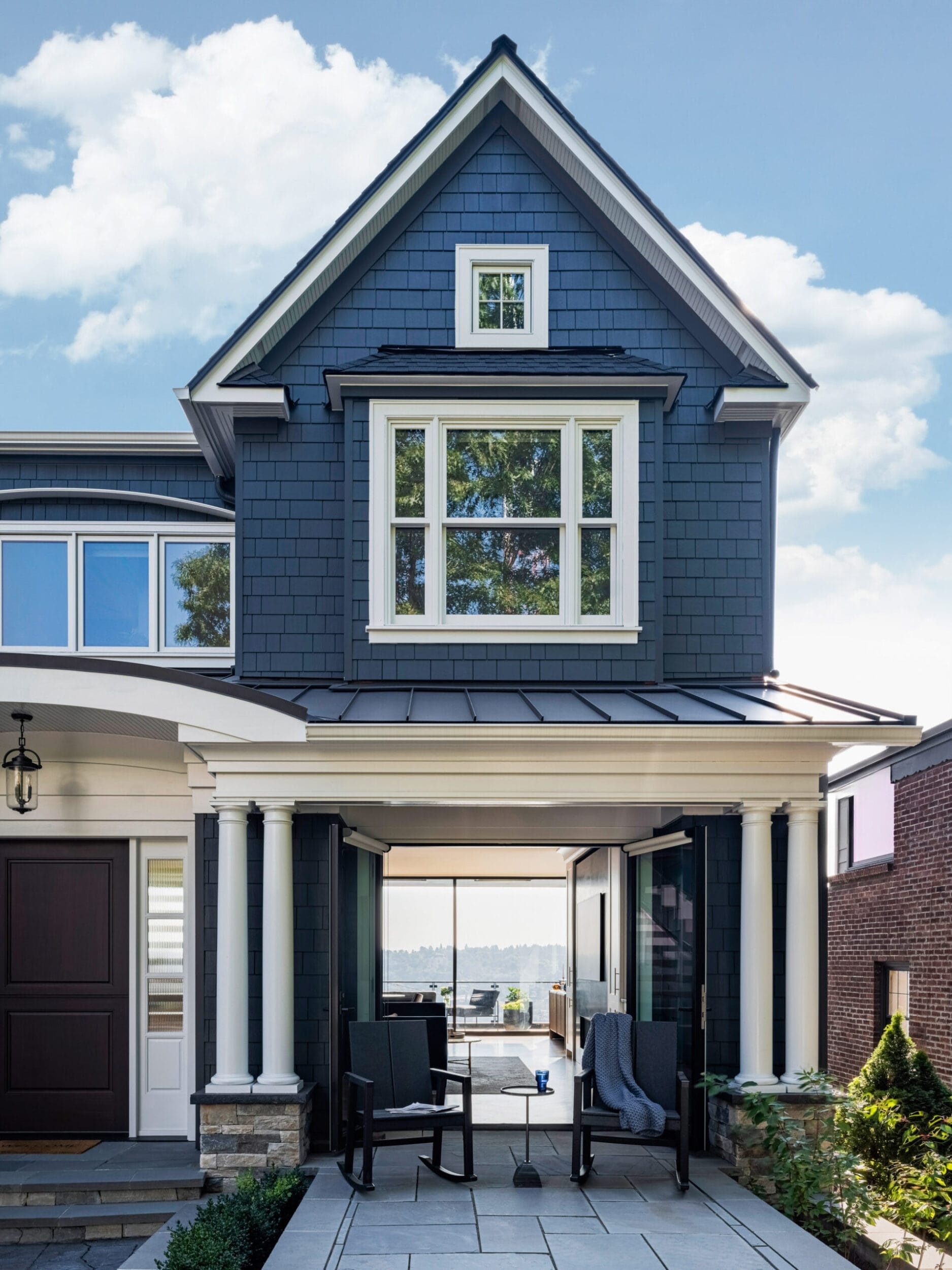 A two-story house with dark blue shingles and white trim. The entrance features columns and outdoor seating on a patio. A view through the house shows a body of water in the background.