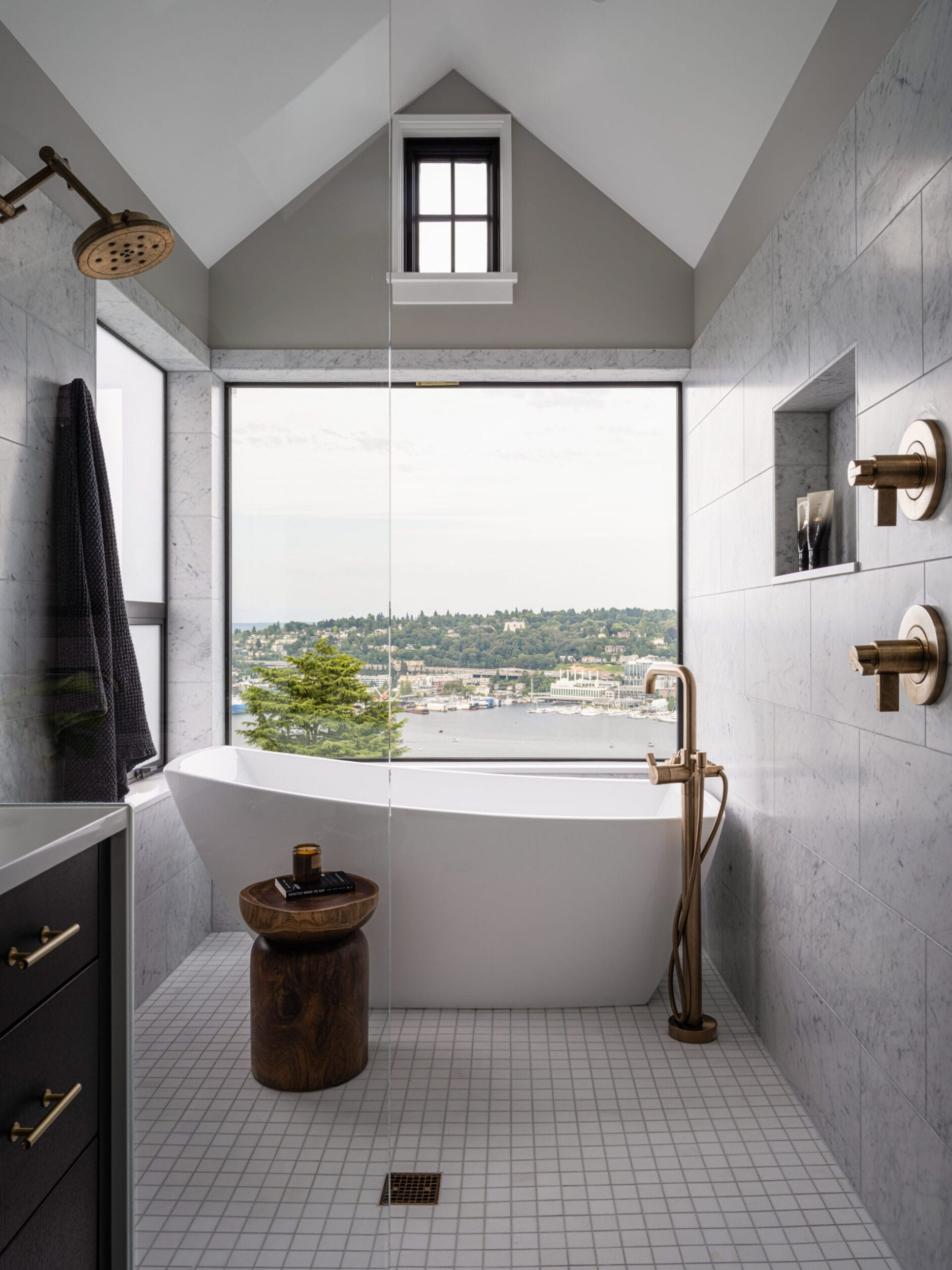 Modern bathroom with a freestanding tub, floor-to-ceiling window, brass fixtures, and a view of a lake. Marble walls and white tile flooring complement the rustic wooden stool.