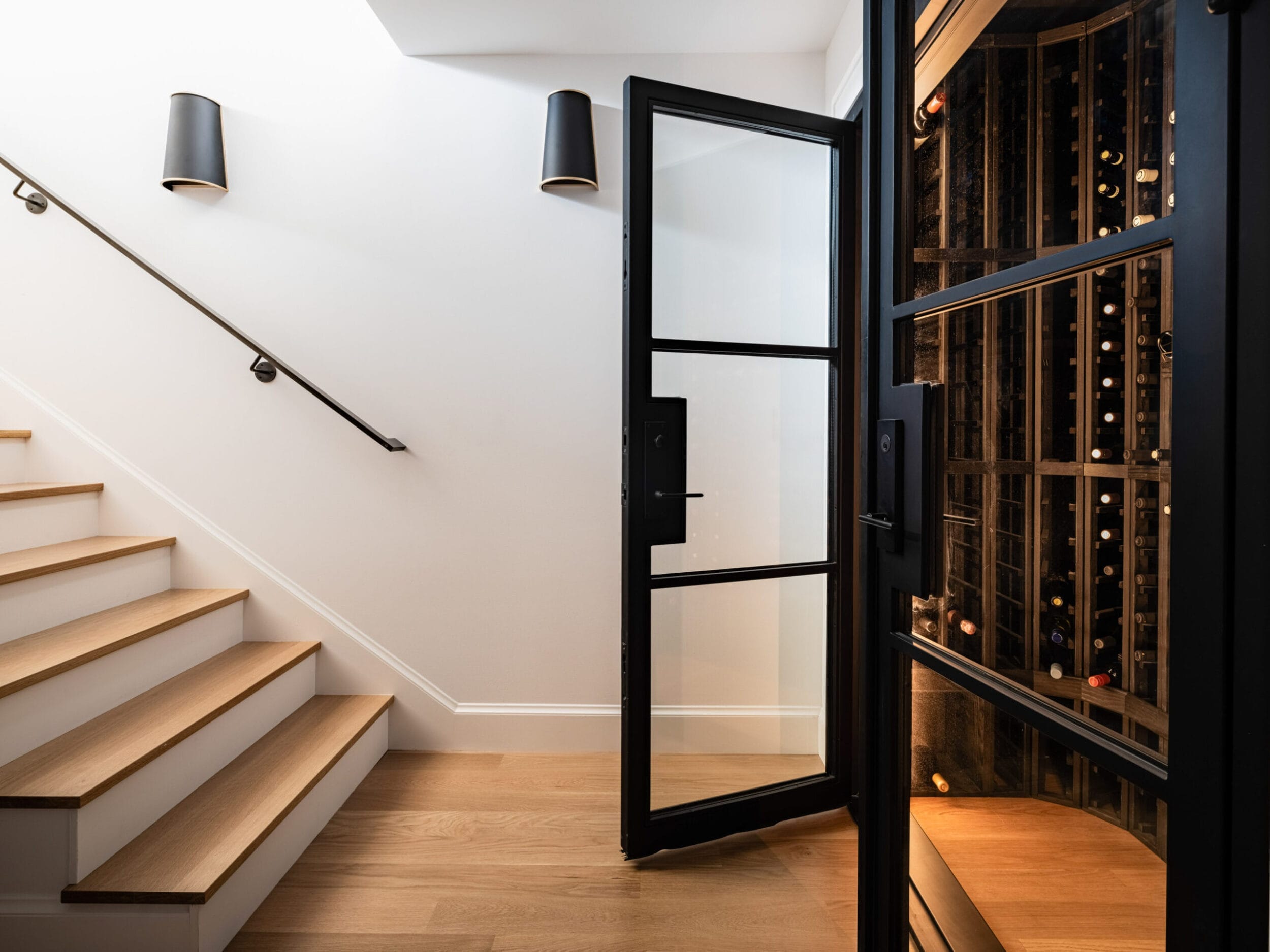 Modern staircase next to a wine cellar with a glass door. The cellar is lit and stocked with wine bottles.