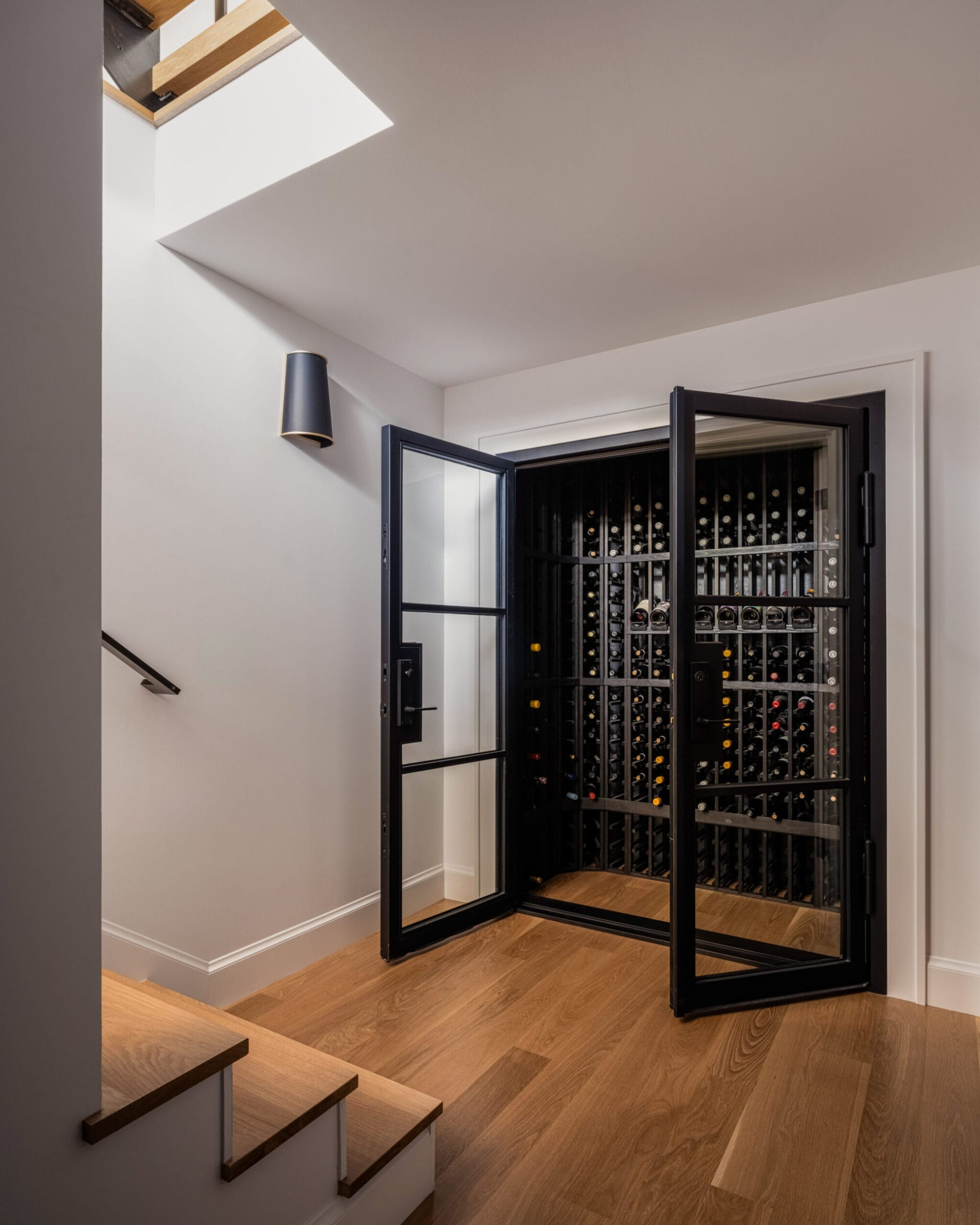 A wine cellar with an open glass door, featuring rows of wine bottles on shelves, located at the bottom of a staircase with wooden steps and a skylight above.