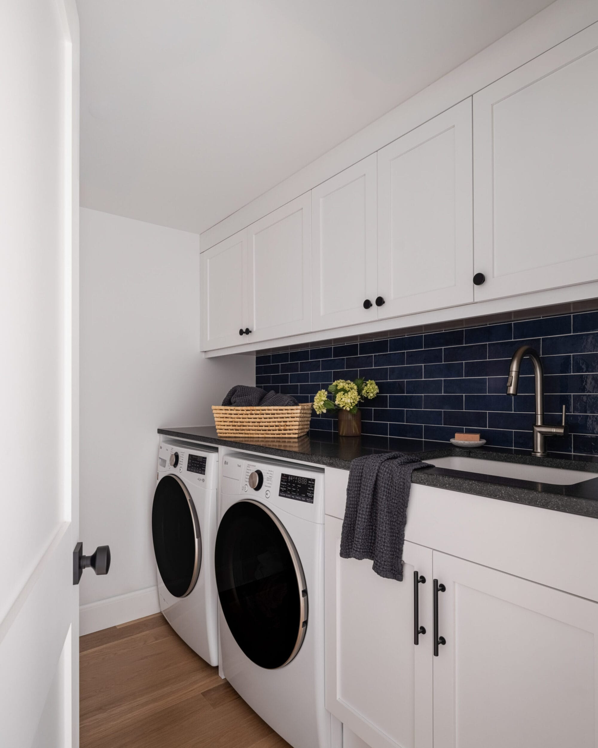 Laundry room with white cabinets, a black countertop, and a stainless steel sink. A washer and dryer are side by side with a basket and flowers on top. Black tile backsplash.