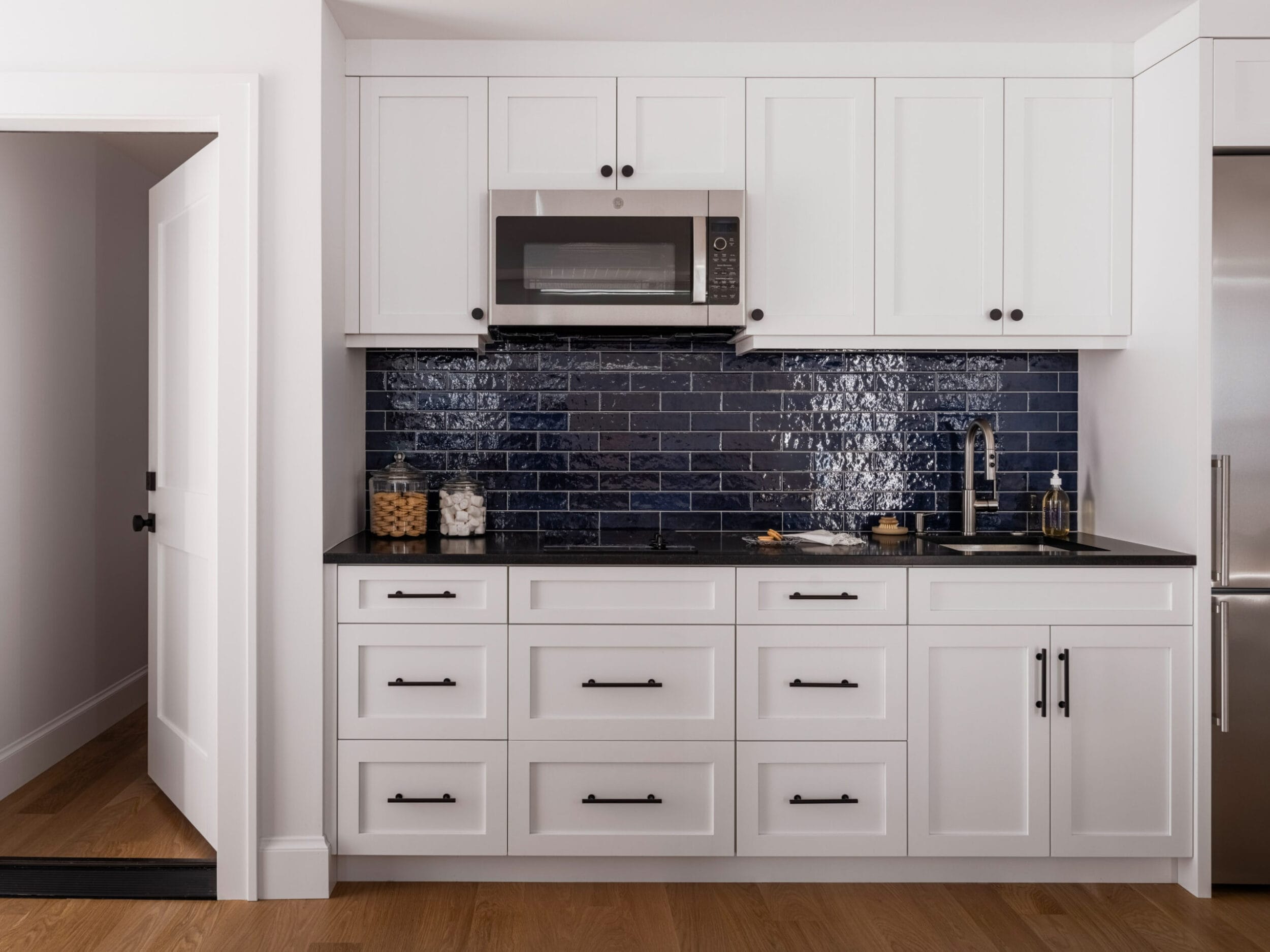 A modern kitchen with white cabinets, a dark blue tile backsplash, black countertop, a microwave, and wood flooring.