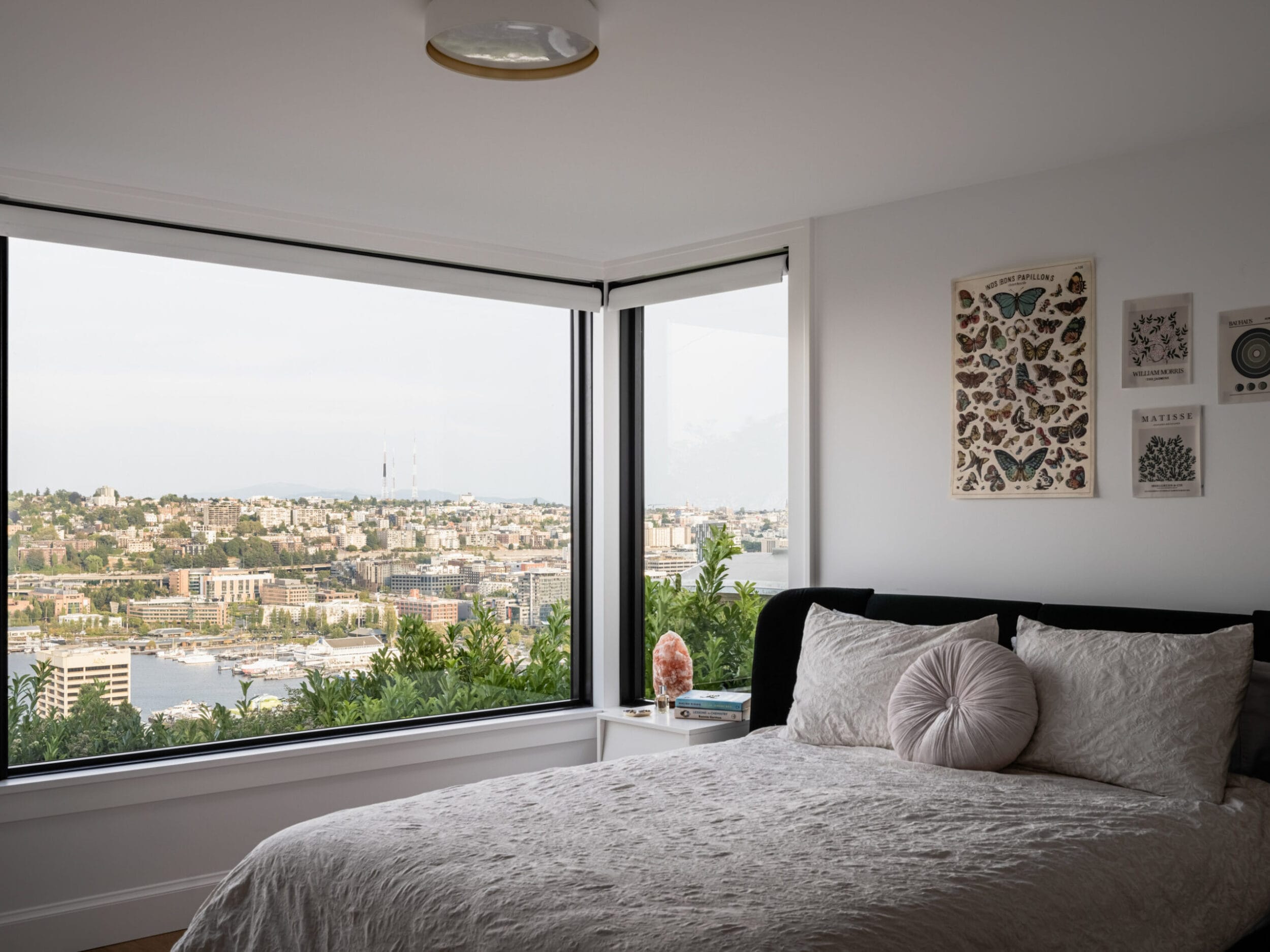 A bedroom with a double bed, decorative pillows, and art on the walls overlooks a cityscape through large corner windows.