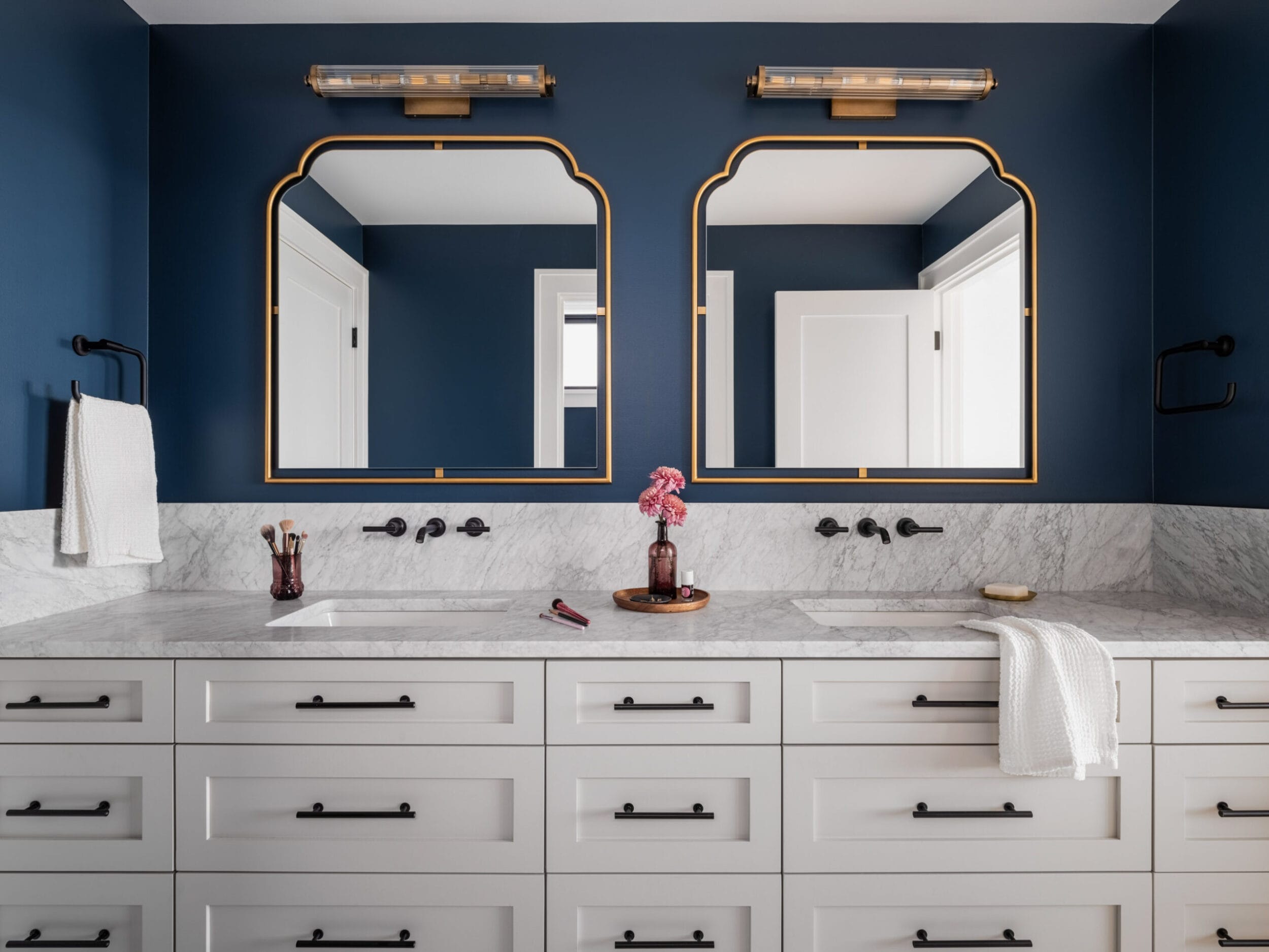 A bathroom with a double vanity, marble countertop, two mirrors, dark blue walls, and white cabinets. A vase with pink flowers and toiletries are placed on the counter.