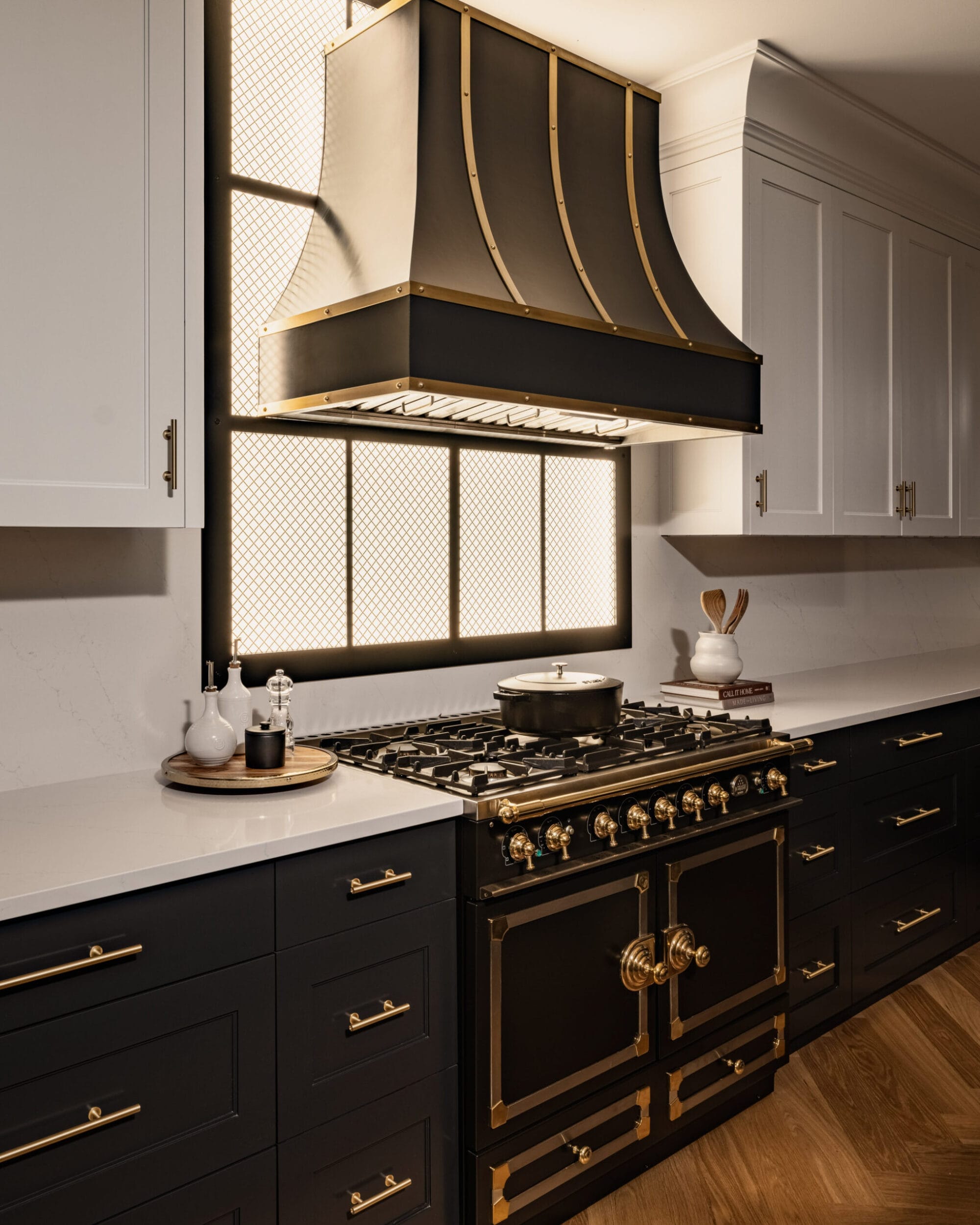 A modern kitchen with a black and gold stove, black lower cabinets, white upper cabinets, and a large black range hood against a backlit grid window.