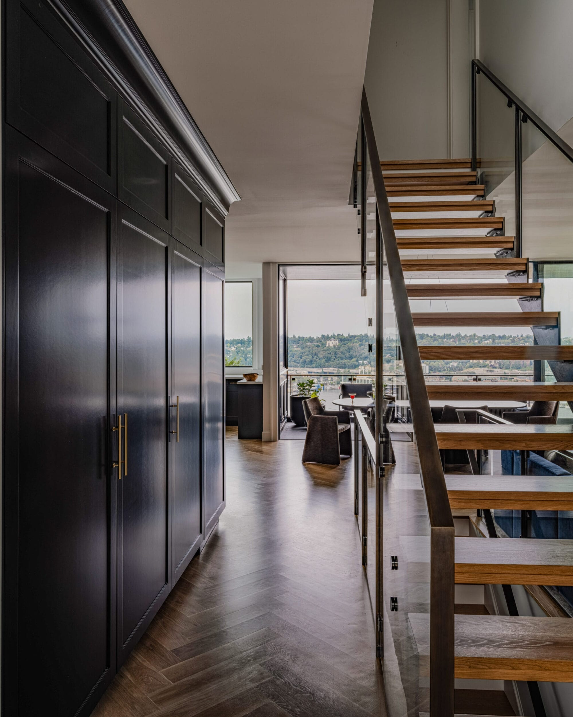 Modern interior with a wooden staircase on the right, dark cabinets on the left, and large windows offering a view of greenery in the background.